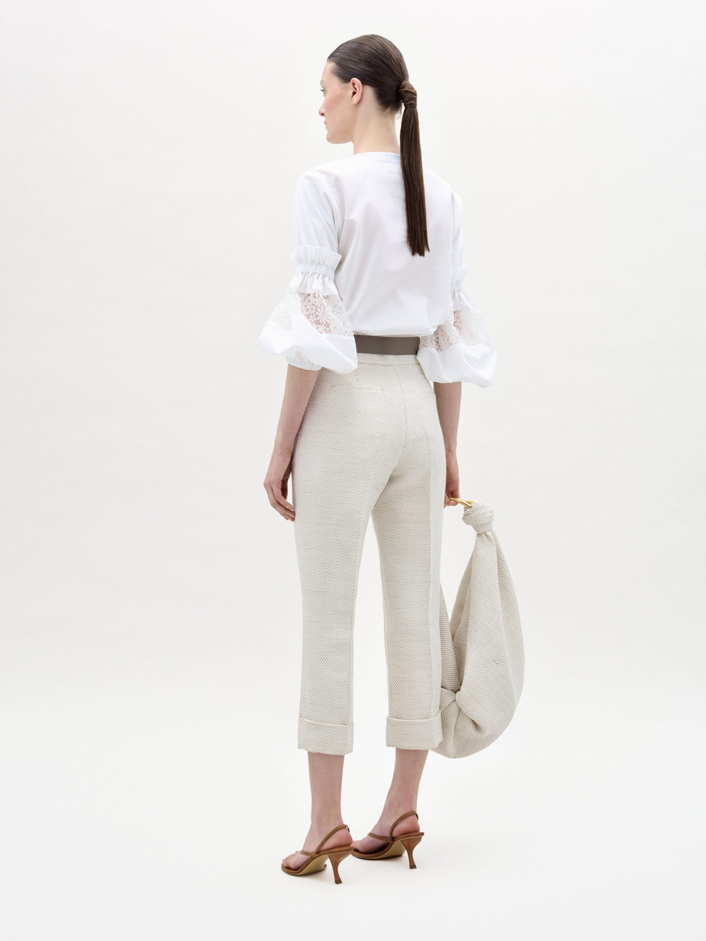 A woman faces away, wearing SVT DESIGNS’ Moad Pant—high-waisted, straight cropped leg in luxe Italian fabric—with a white lace ruffle sleeve blouse. She holds a light bag, hair in a low ponytail, set against a plain background.