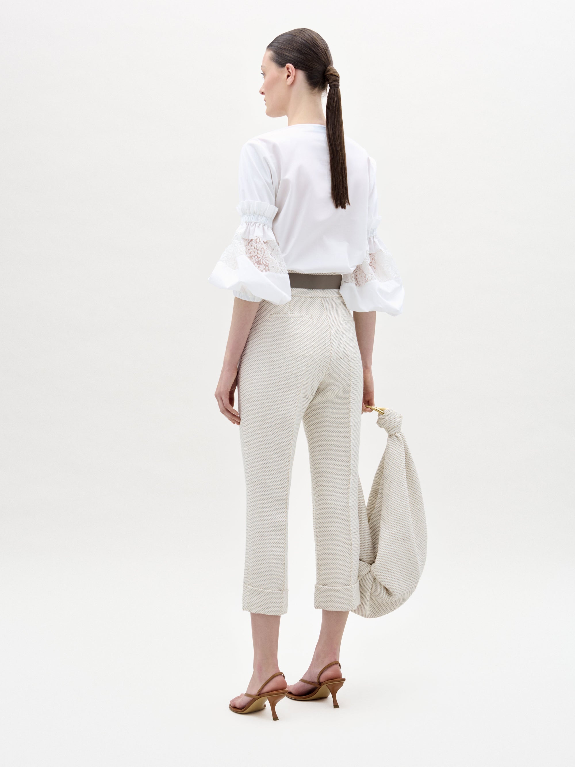 A woman faces away, wearing SVT DESIGNS’ Moad Pant—high-waisted, straight cropped leg in luxe Italian fabric—with a white lace ruffle sleeve blouse. She holds a light bag, hair in a low ponytail, set against a plain background.