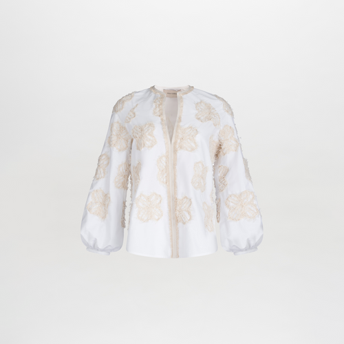 The Molveno Blouse by SVT DESIGNS is a white embroidered cotton piece with a round neckline. It features sheer fabric with beige floral motifs throughout, offering a refined look against a plain white backdrop.