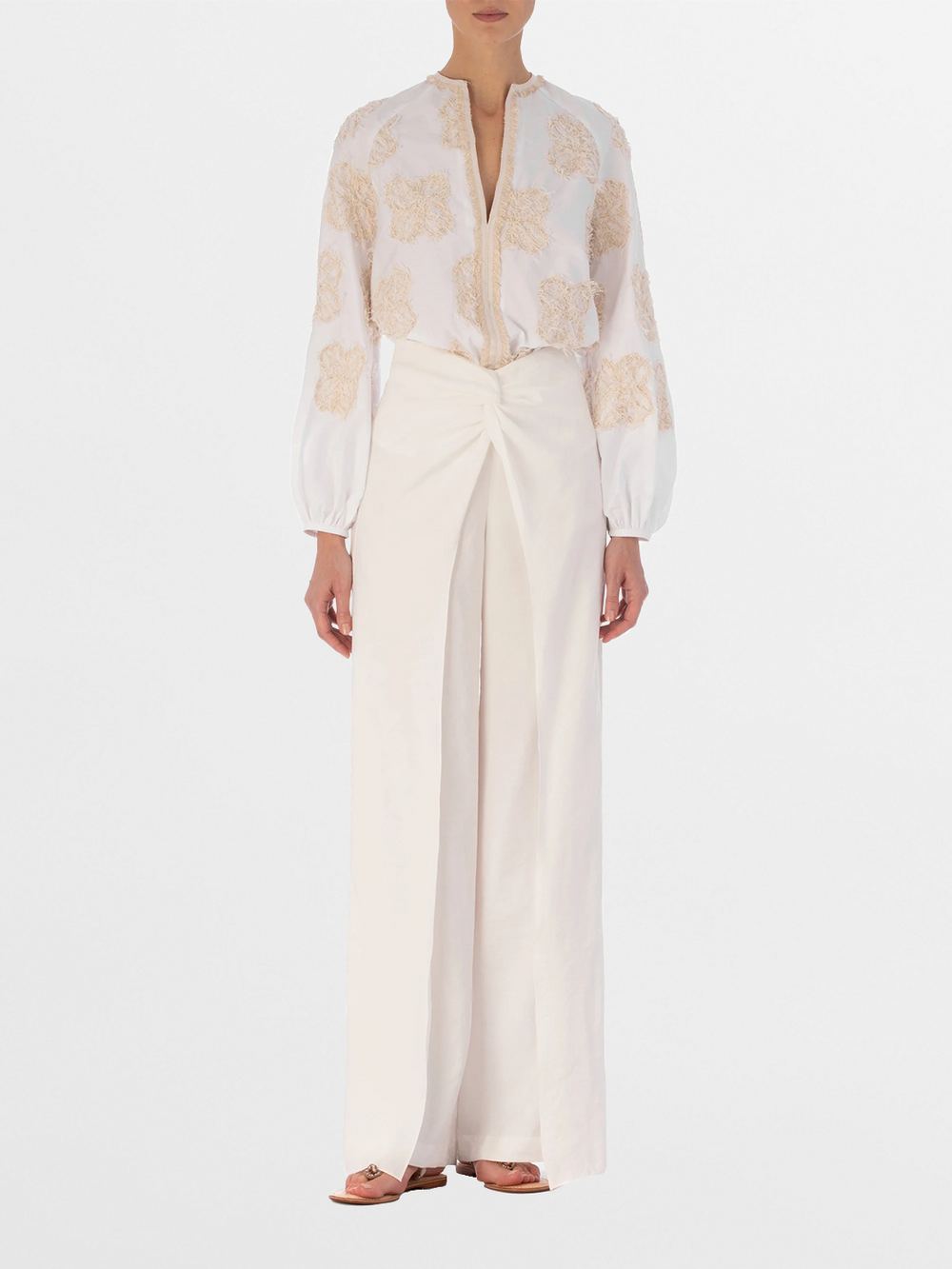 A person wearing the SVT DESIGNS Molveno Blouse—white cotton with beige floral embroidery and long sleeves—paired with high-waisted, wide-leg white pants and flat sandals, against a plain white background.