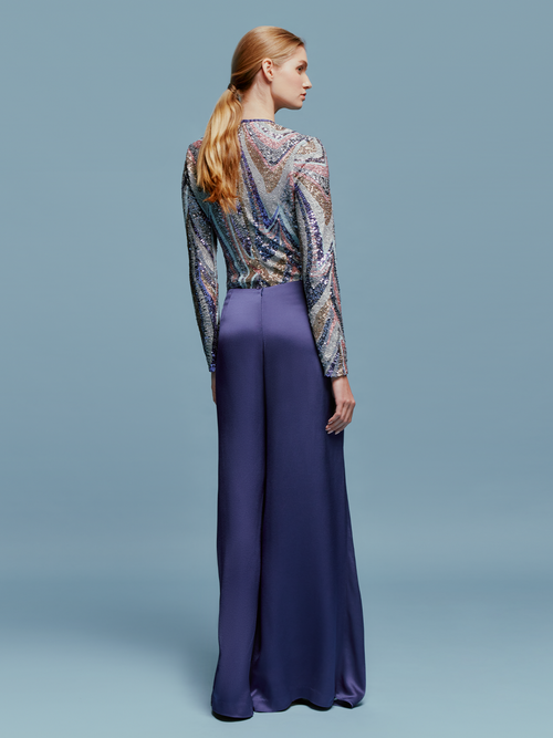 A person stands against a light blue background wearing a patterned long-sleeve top and the high-waisted, wide-leg Nadira Pant in Navy, ready for shipping by August 2024.