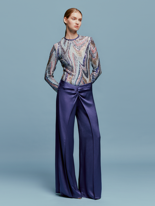 A person stands against a light blue background wearing a patterned long-sleeve top and the high-waisted, wide-leg Nadira Pant in Navy, ready for shipping by August 2024.