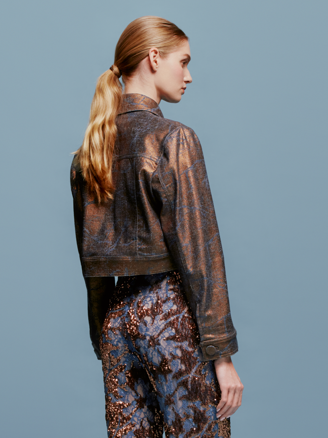 Against a blue background, a woman with light brown hair in a low ponytail wears the Nahid Jacket Metallic Ocher and a blue and brown sequin skirt. The scene captures the essence of chic autumn fashion, as if heralding the trends for September 2nd, 2024.