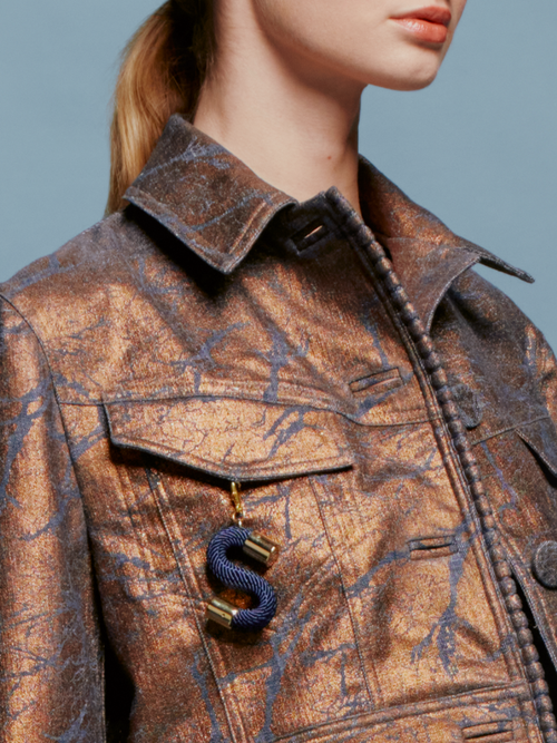 Against a blue background, a woman with light brown hair in a low ponytail wears the Nahid Jacket Metallic Ocher and a blue and brown sequin skirt. The scene captures the essence of chic autumn fashion, as if heralding the trends for September 2nd, 2024.