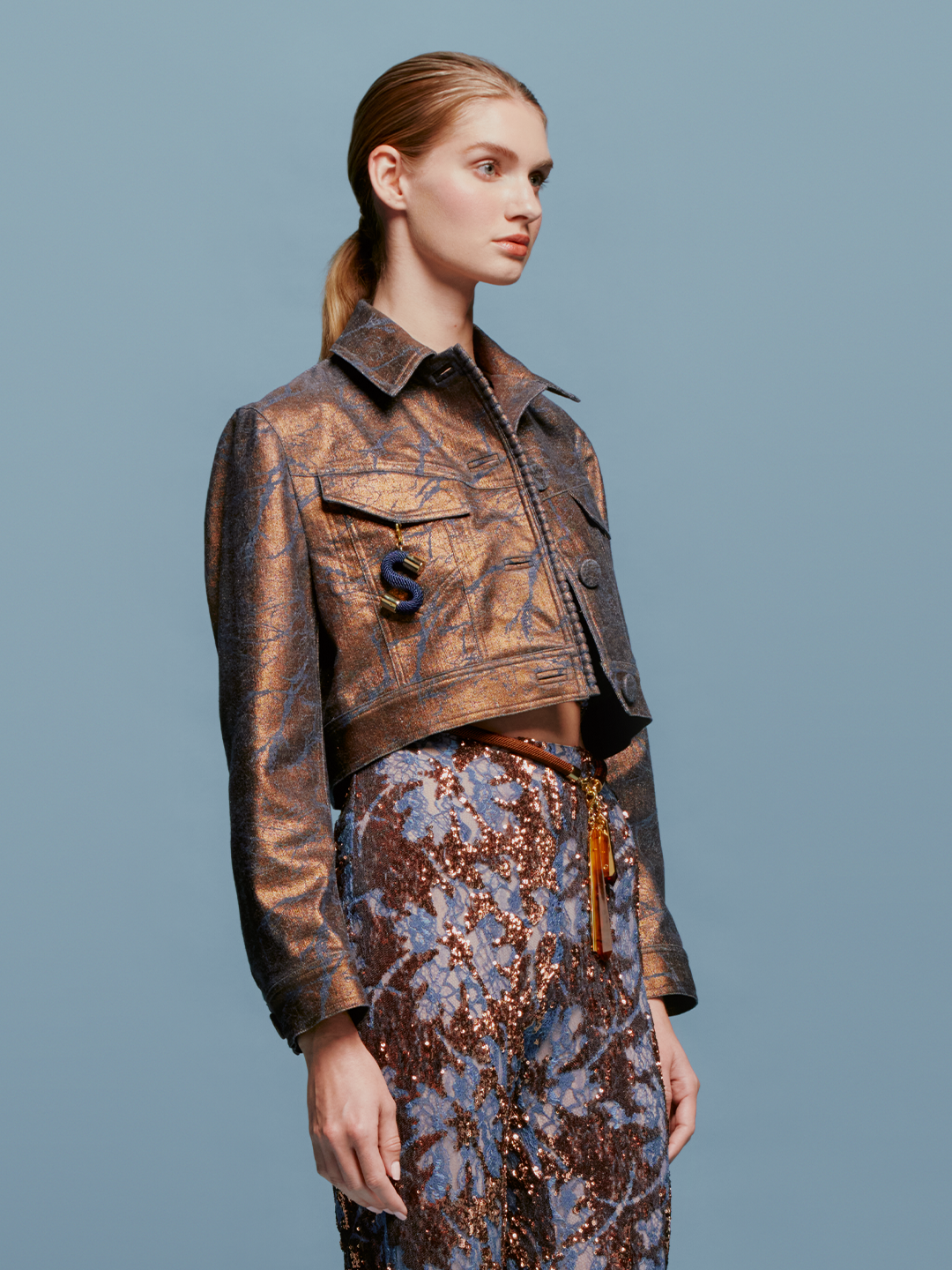 Against a blue background, a woman with light brown hair in a low ponytail wears the Nahid Jacket Metallic Ocher and a blue and brown sequin skirt. The scene captures the essence of chic autumn fashion, as if heralding the trends for September 2nd, 2024.