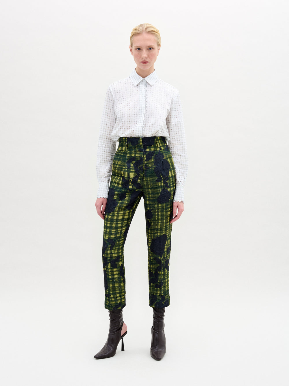 Against a white backdrop, a person confidently wears SVT DESIGNS' Naroa Blouse made of Italian fabric with intricate patterns. They pair it with abstract green and black plaid pants and black high-heeled boots. They have short blonde hair and meet the camera's gaze confidently.