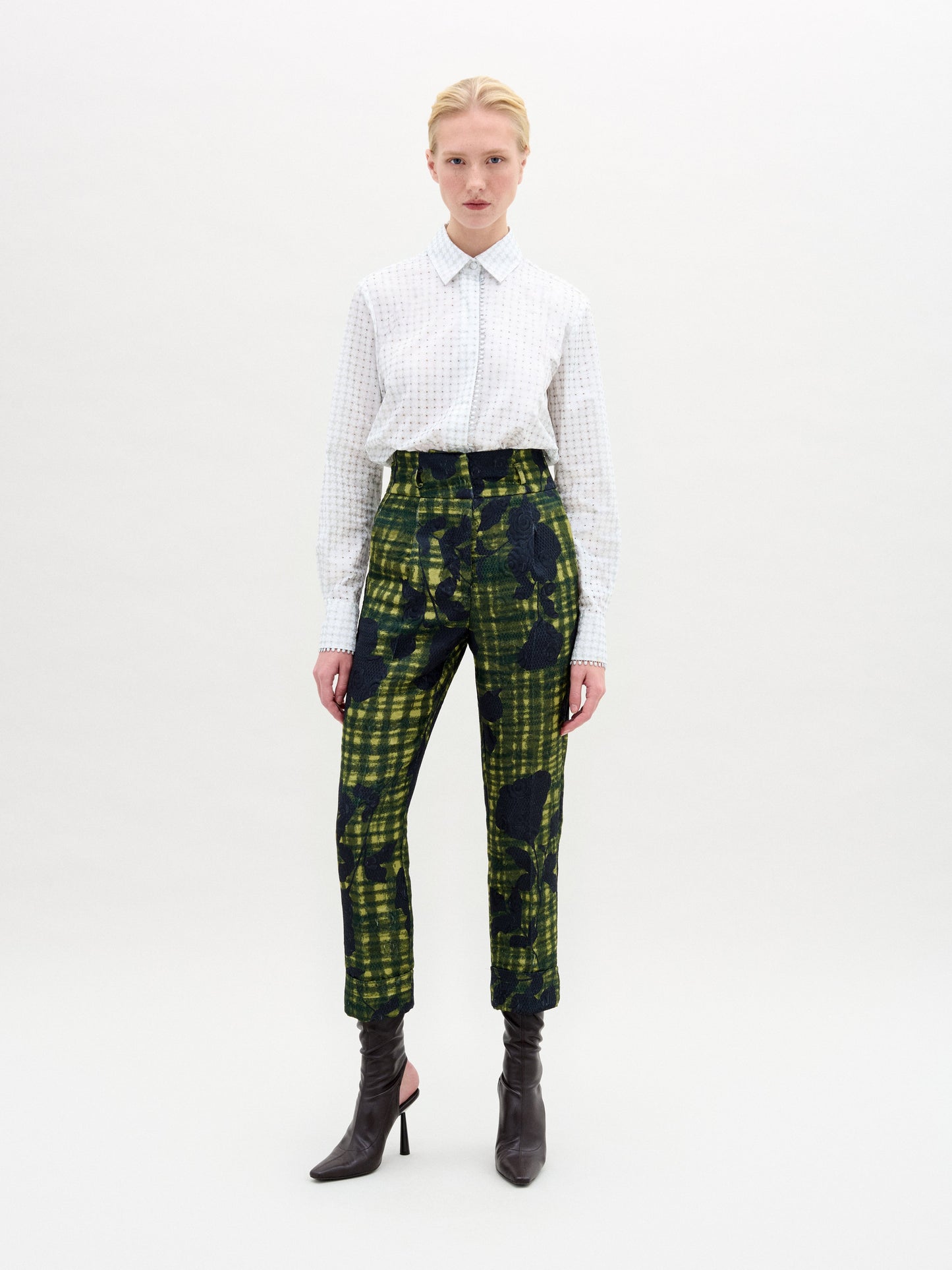 Against a white backdrop, a person confidently wears SVT DESIGNS' Naroa Blouse made of Italian fabric with intricate patterns. They pair it with abstract green and black plaid pants and black high-heeled boots. They have short blonde hair and meet the camera's gaze confidently.