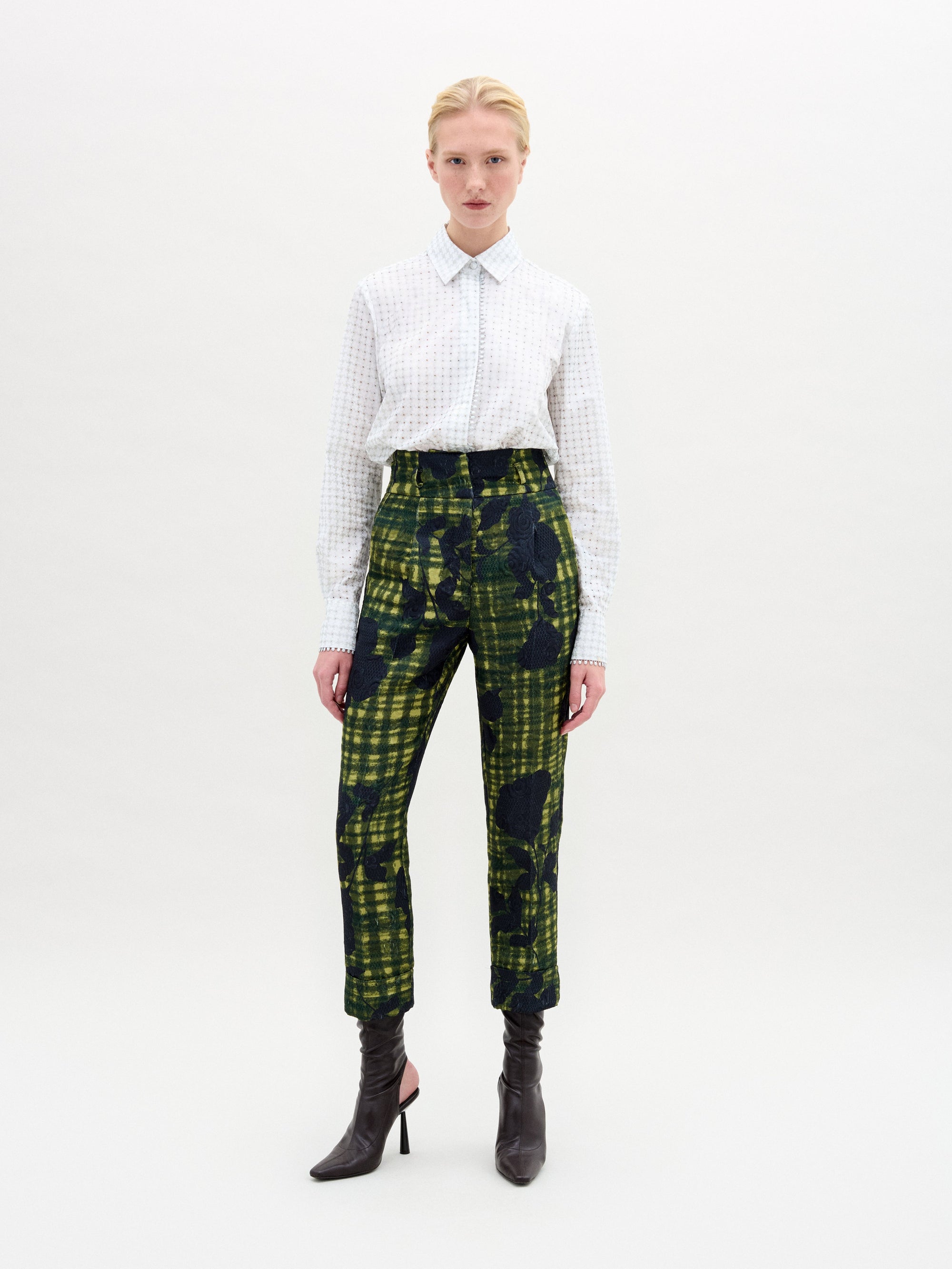 Against a white backdrop, a person confidently wears SVT DESIGNS' Naroa Blouse made of Italian fabric with intricate patterns. They pair it with abstract green and black plaid pants and black high-heeled boots. They have short blonde hair and meet the camera's gaze confidently.