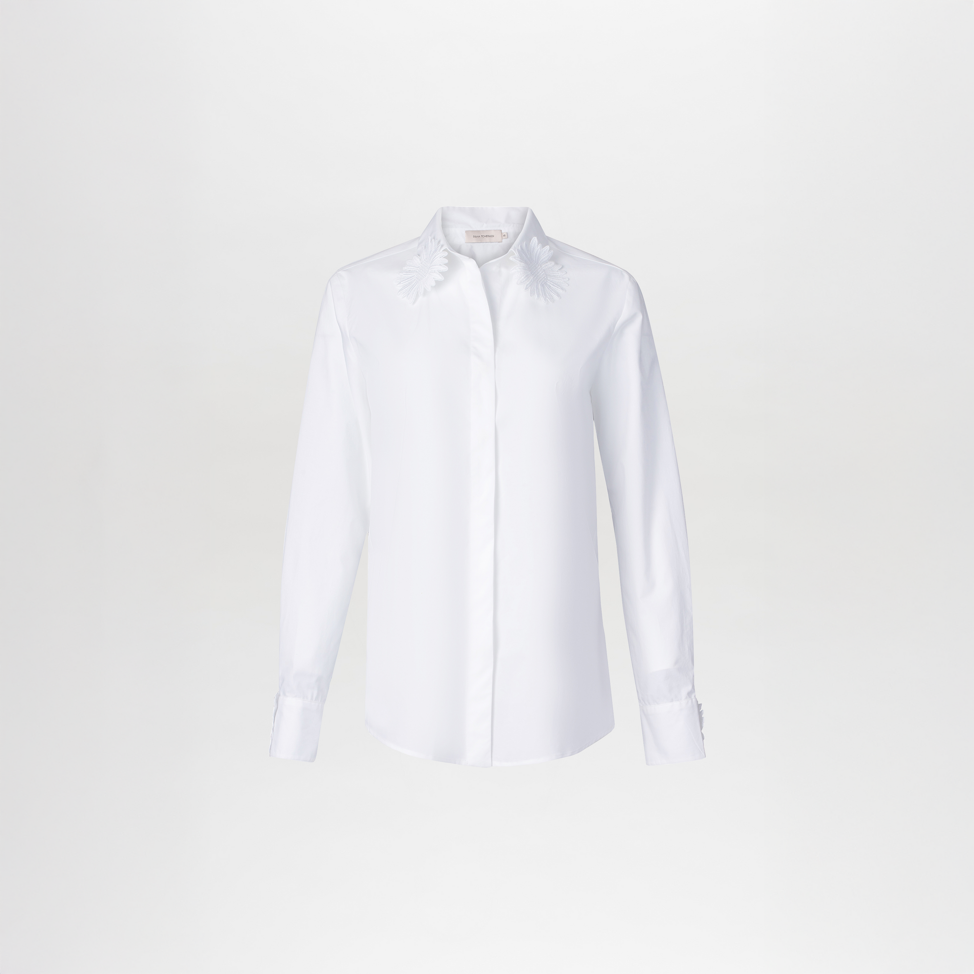The Naroa Blouse by SVT DESIGNS features a crisp white cotton fabric with floral embroidery on each collar tip, complemented by a traditional shirt collar and classic button-down front with buttoned cuffs.