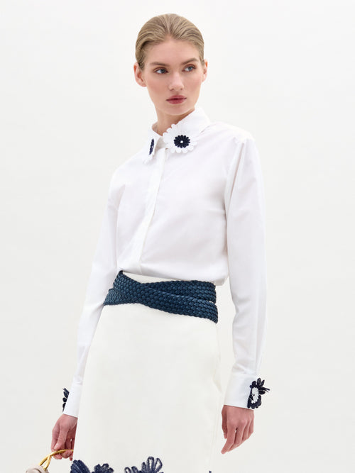 A person models the SVT DESIGNS Naroa Blouse, a white button-down with floral embroidery on the collar and cuffs. It's paired with a skirt featuring appliqué details near the hem, cinched by a dark woven belt of fine Italian fabric, set against a plain background.