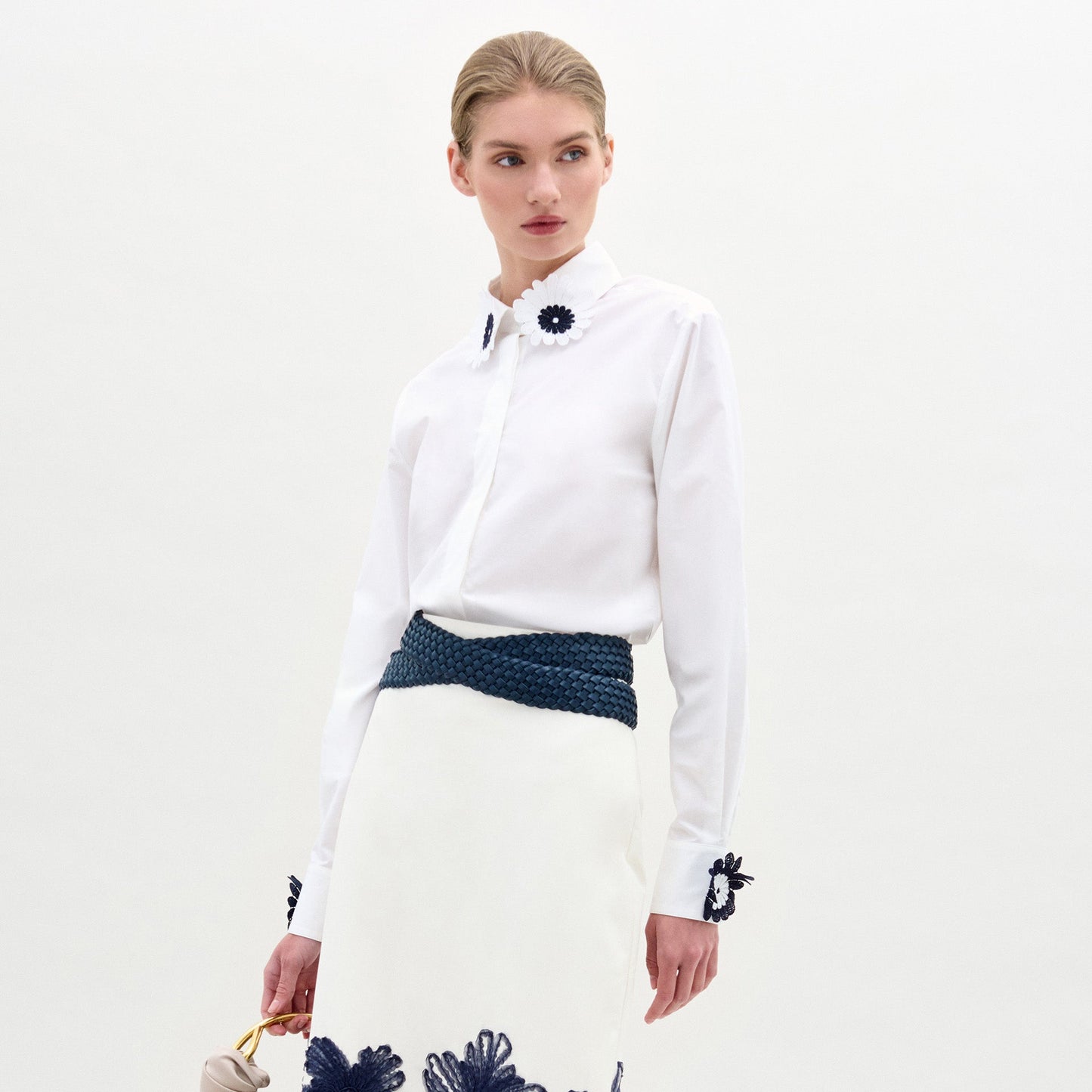 A woman stands against a plain white background wearing the Naroa Blouse by SVT DESIGNS, a long-sleeve Italian-fabric top with floral appliqué, paired with a white skirt with blue floral accents, a braided blue belt, and holding a light handbag.