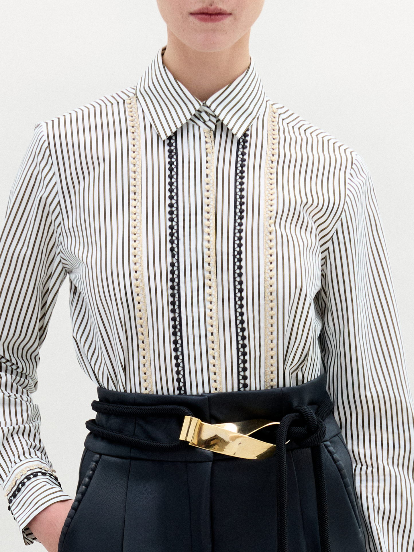 Person stands against a plain background, wearing the Naroa Blouse— a striped 100% cotton button-up— tucked into high-waisted black pants with a gold-buckle belt.