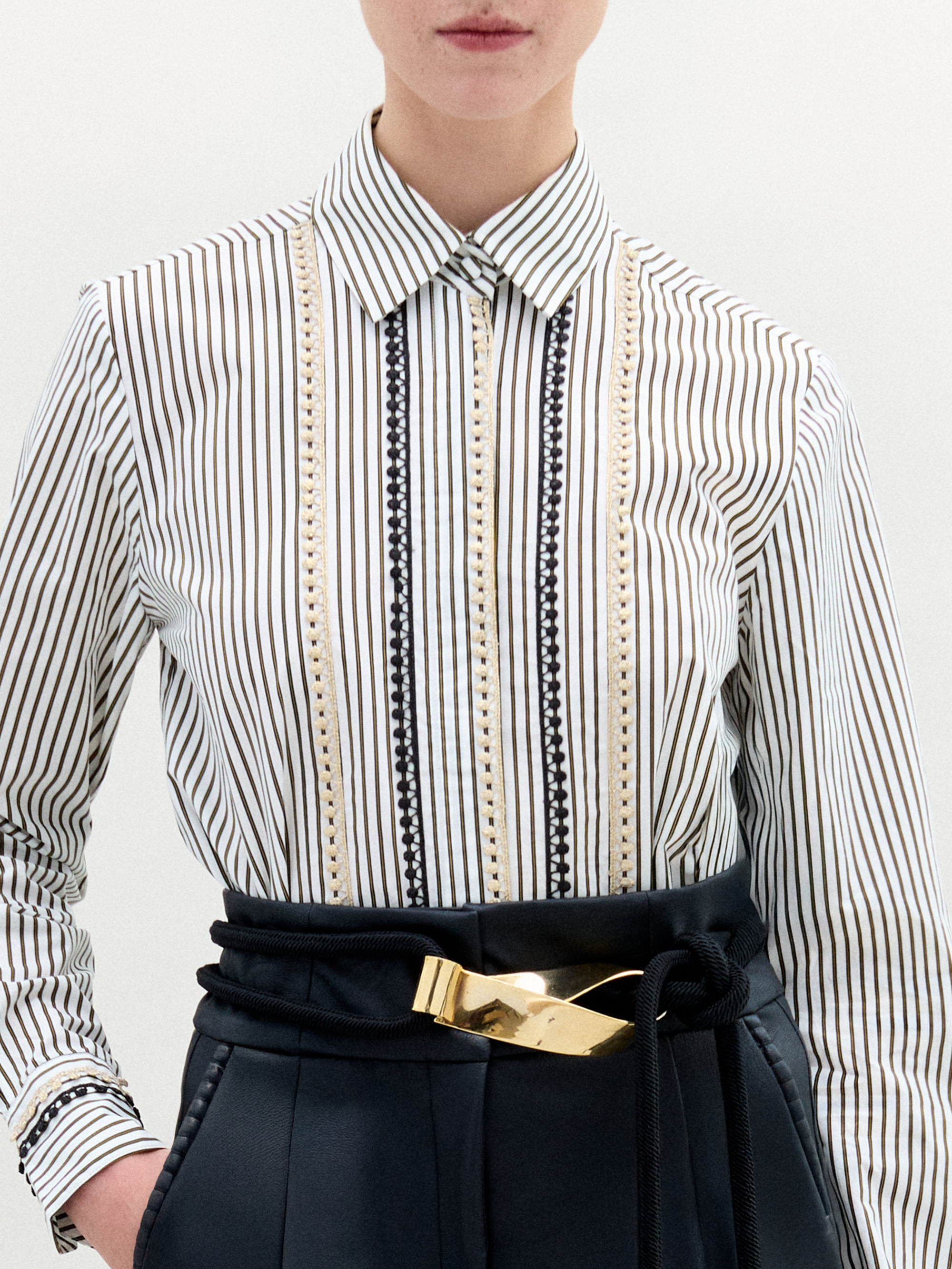 Person stands against a plain background, wearing the Naroa Blouse— a striped 100% cotton button-up— tucked into high-waisted black pants with a gold-buckle belt.