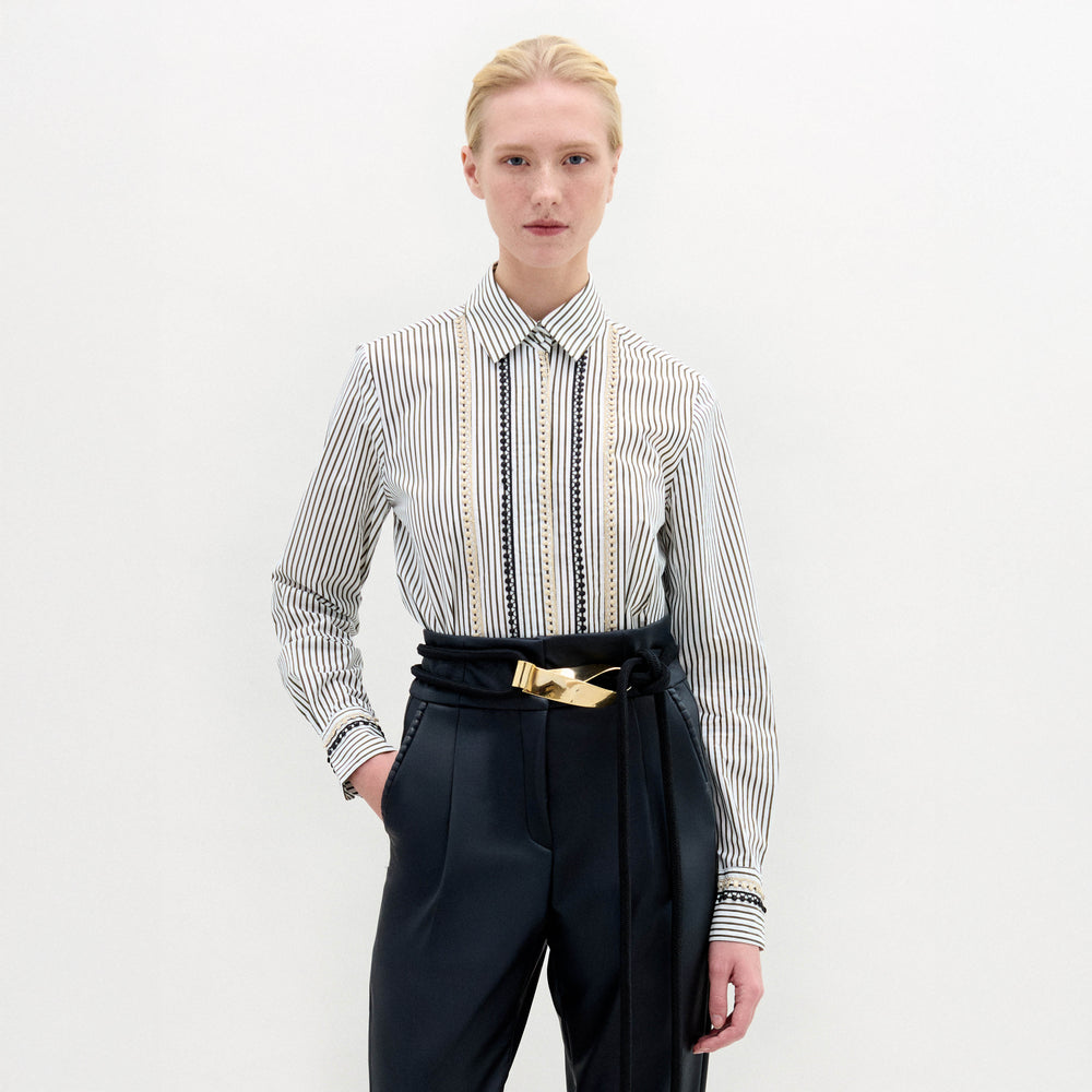 A person with pulled-back blonde hair and a neutral expression wears the SVT DESIGNS Naroa Blouse—striped, long-sleeve, 100% cotton Italian fabric—tucked into high-waisted black trousers with a gold-buckle belt against a white background.