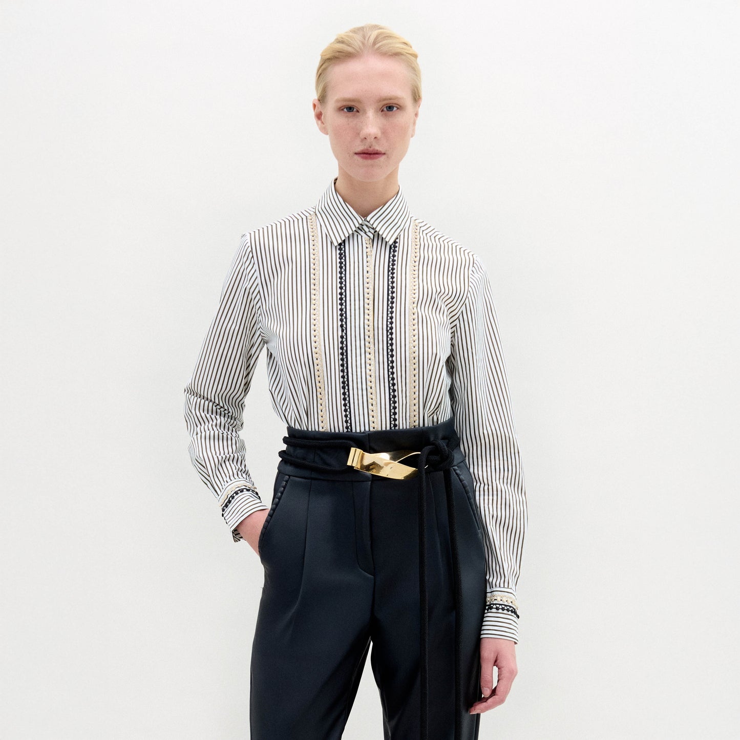 A person with pulled-back blonde hair and a neutral expression wears the SVT DESIGNS Naroa Blouse—striped, long-sleeve, 100% cotton Italian fabric—tucked into high-waisted black trousers with a gold-buckle belt against a white background.