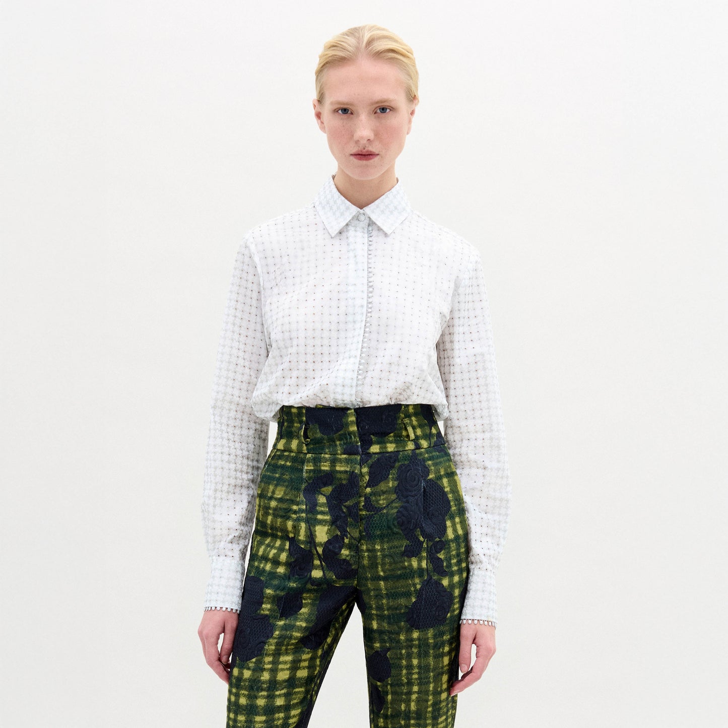 Against a neutral backdrop, a person models a chic outfit with the Naroa Blouse in Italian white patterned fabric, styled with green and blue patterned pants. The ensemble is completed by sleek black high-heeled boots.