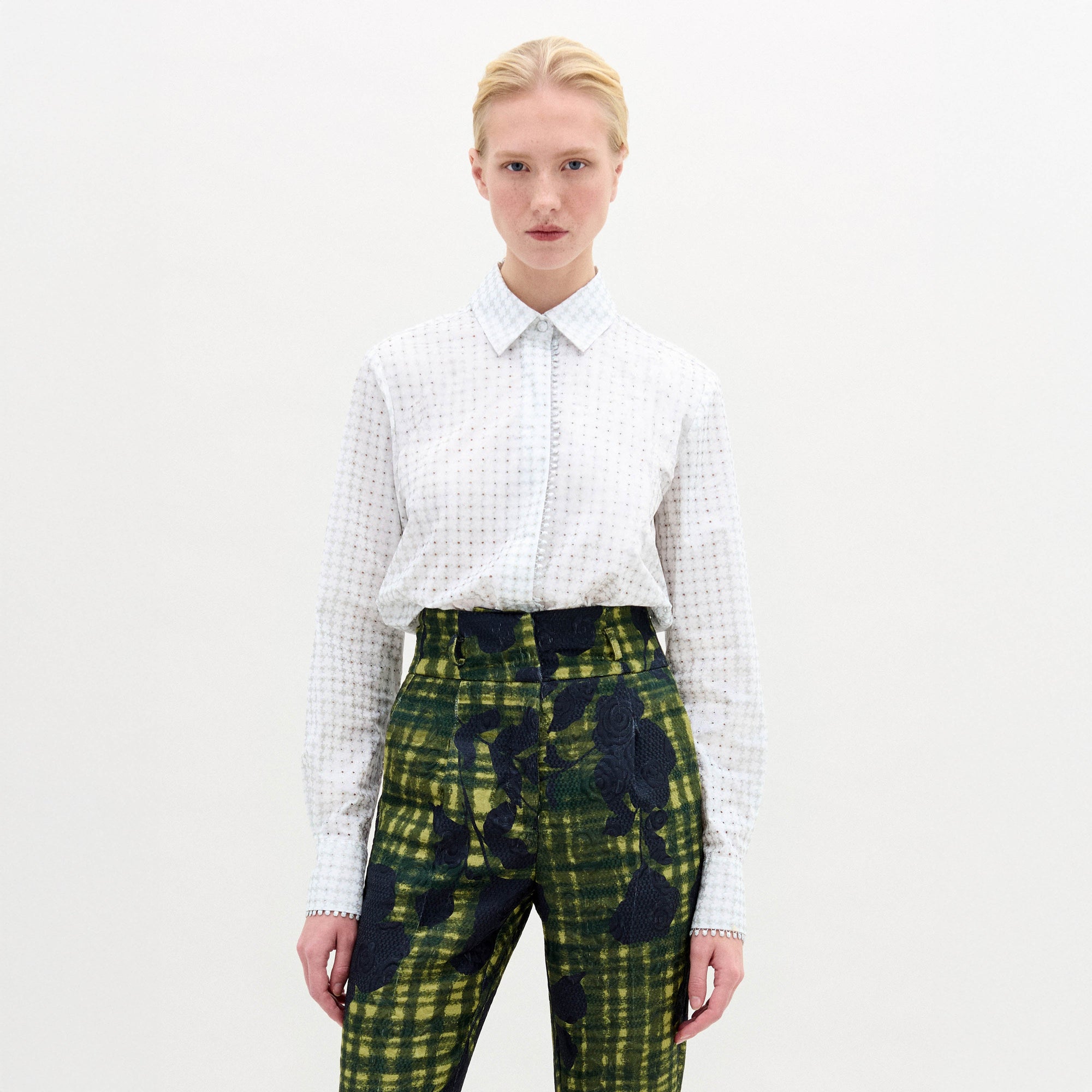 Against a neutral backdrop, a person models a chic outfit with the Naroa Blouse in Italian white patterned fabric, styled with green and blue patterned pants. The ensemble is completed by sleek black high-heeled boots.