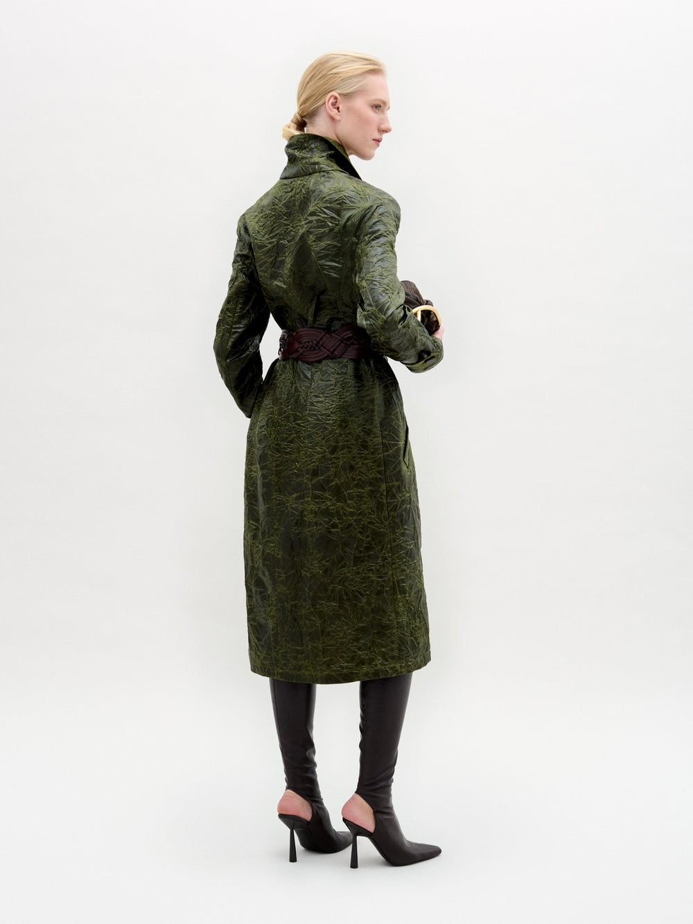 A woman wears the Nilo Trench, a textured dark green coat crafted from Italian fabric, paired with dark boots, a large gold belt buckle, and holding a small dark clutch against a plain background.