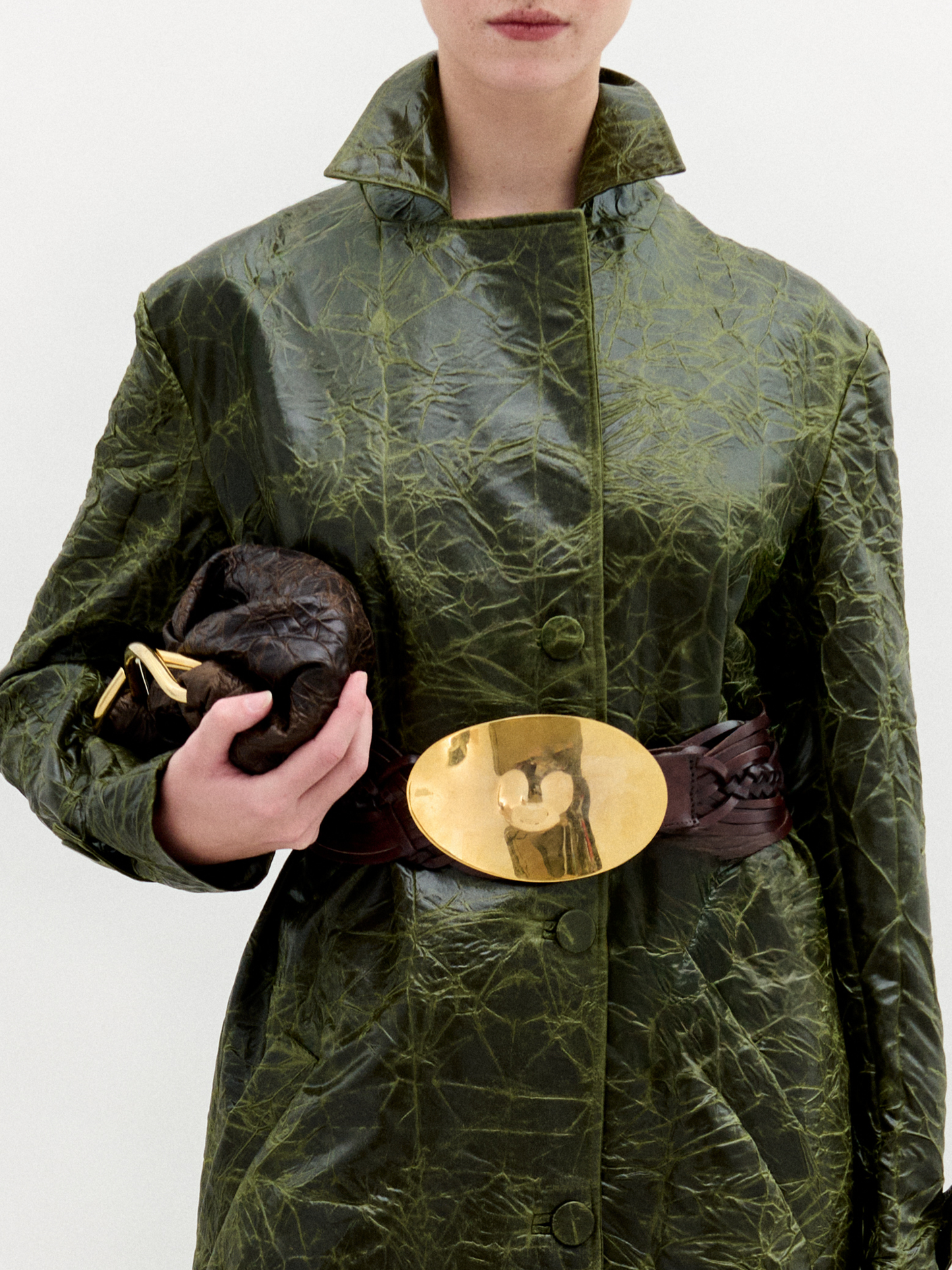 A woman wears the Nilo Trench, a textured dark green coat crafted from Italian fabric, paired with dark boots, a large gold belt buckle, and holding a small dark clutch against a plain background.