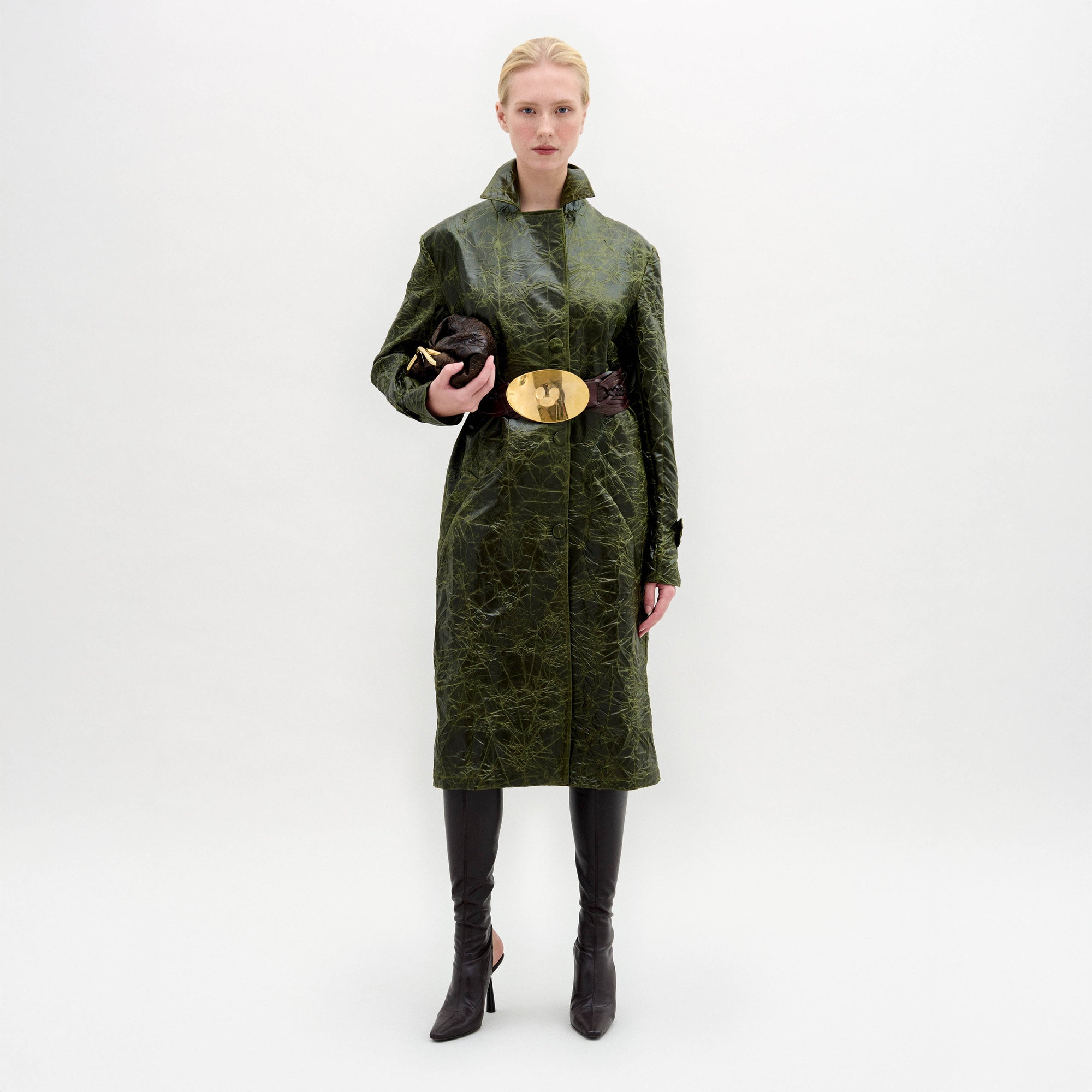 A woman wears the Nilo Trench, a textured dark green coat crafted from Italian fabric, paired with dark boots, a large gold belt buckle, and holding a small dark clutch against a plain background.