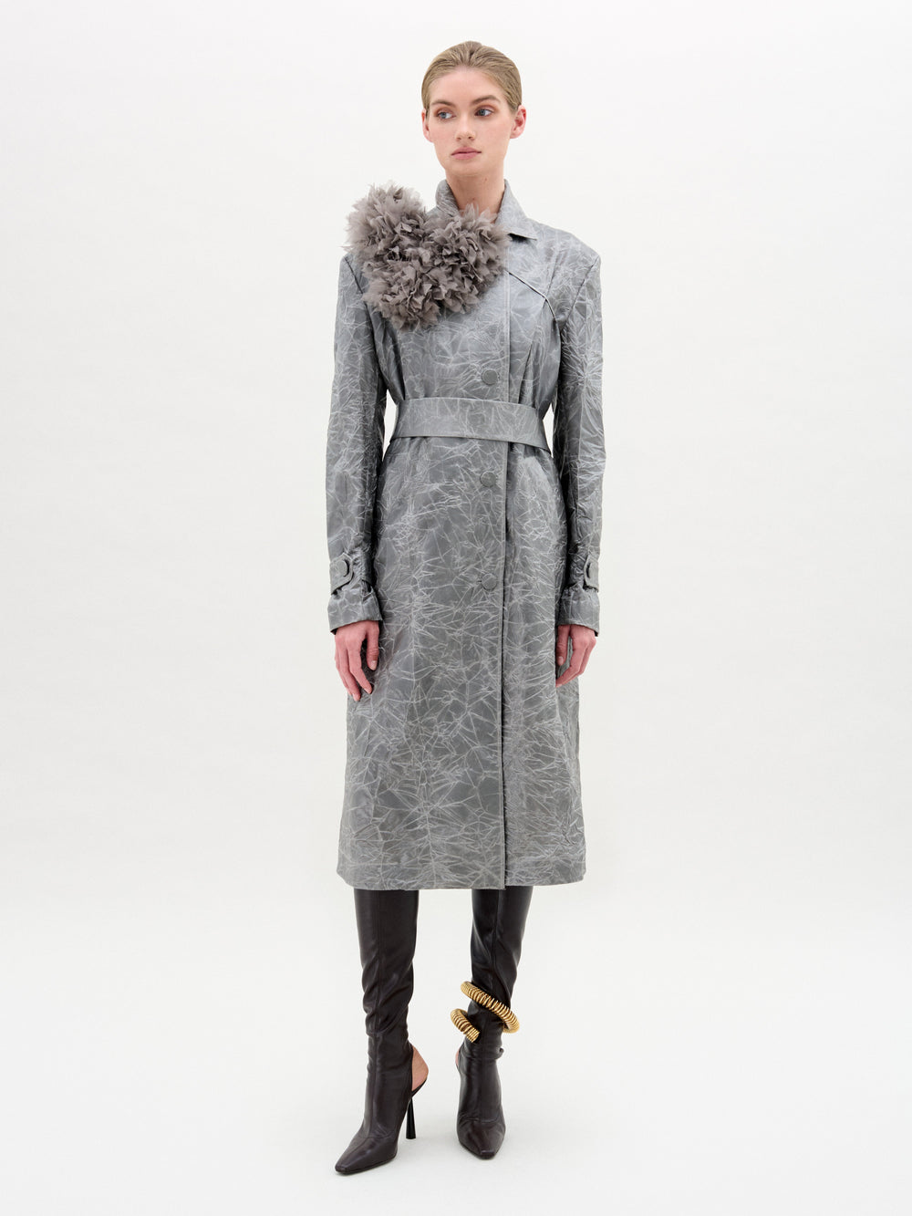 A woman stands against a plain background wearing the Nilo Trench by SVT DESIGNS, a long, textured gray Italian fabric coat with a large fabric flower on the left shoulder, paired with brown tall boots and a chunky gold anklet.