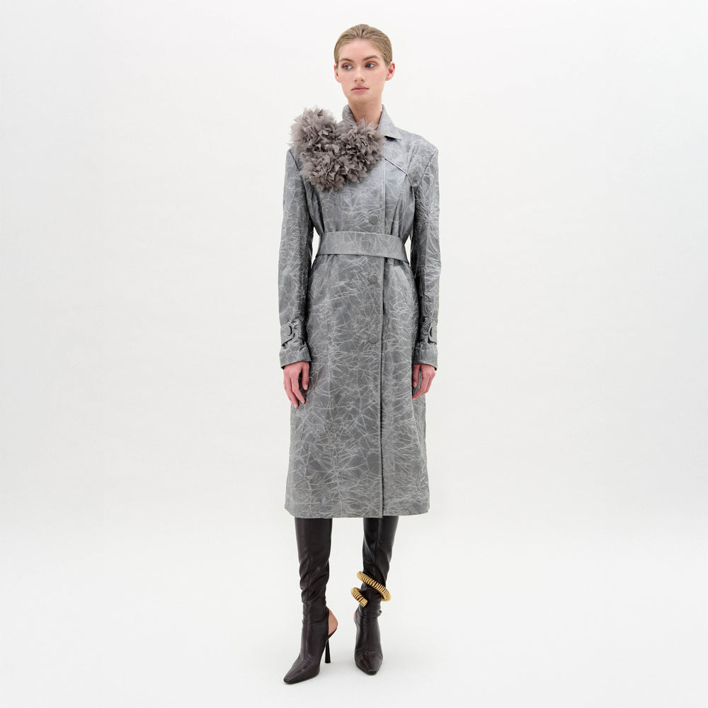 A woman poses against a white background in the Nilo Trench by SVT DESIGNS, a long textured silver Italian trench coat with a large grey floral shoulder detail, paired with black high-heeled boots and gold ankle accessories.