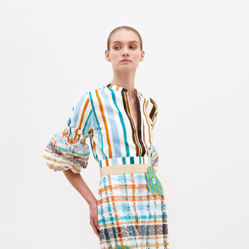 Wearing the Nuru Blouse by SVT DESIGNS, a person stands against a white background. The colorful outfit has blue, orange, and white multi-color stripes with ruffled sleeves, lace details, and a waistband with a circular embellishment.