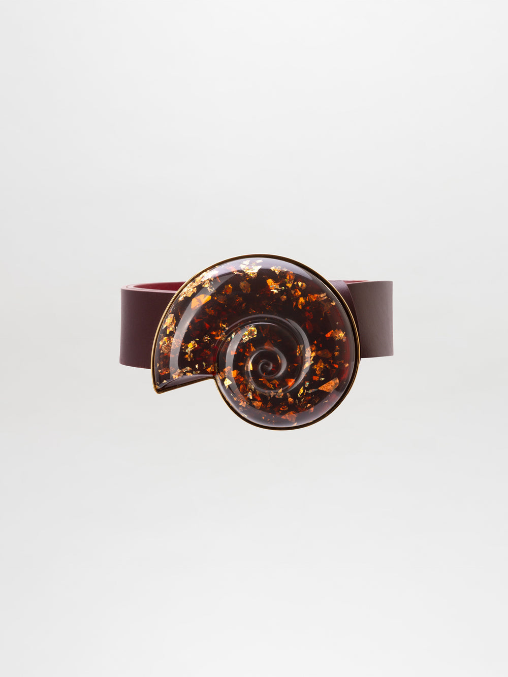 The Nautilus Belt is a dark maroon accessory with a large, glossy, spiral-shaped buckle adorned with amber and gold flecks.