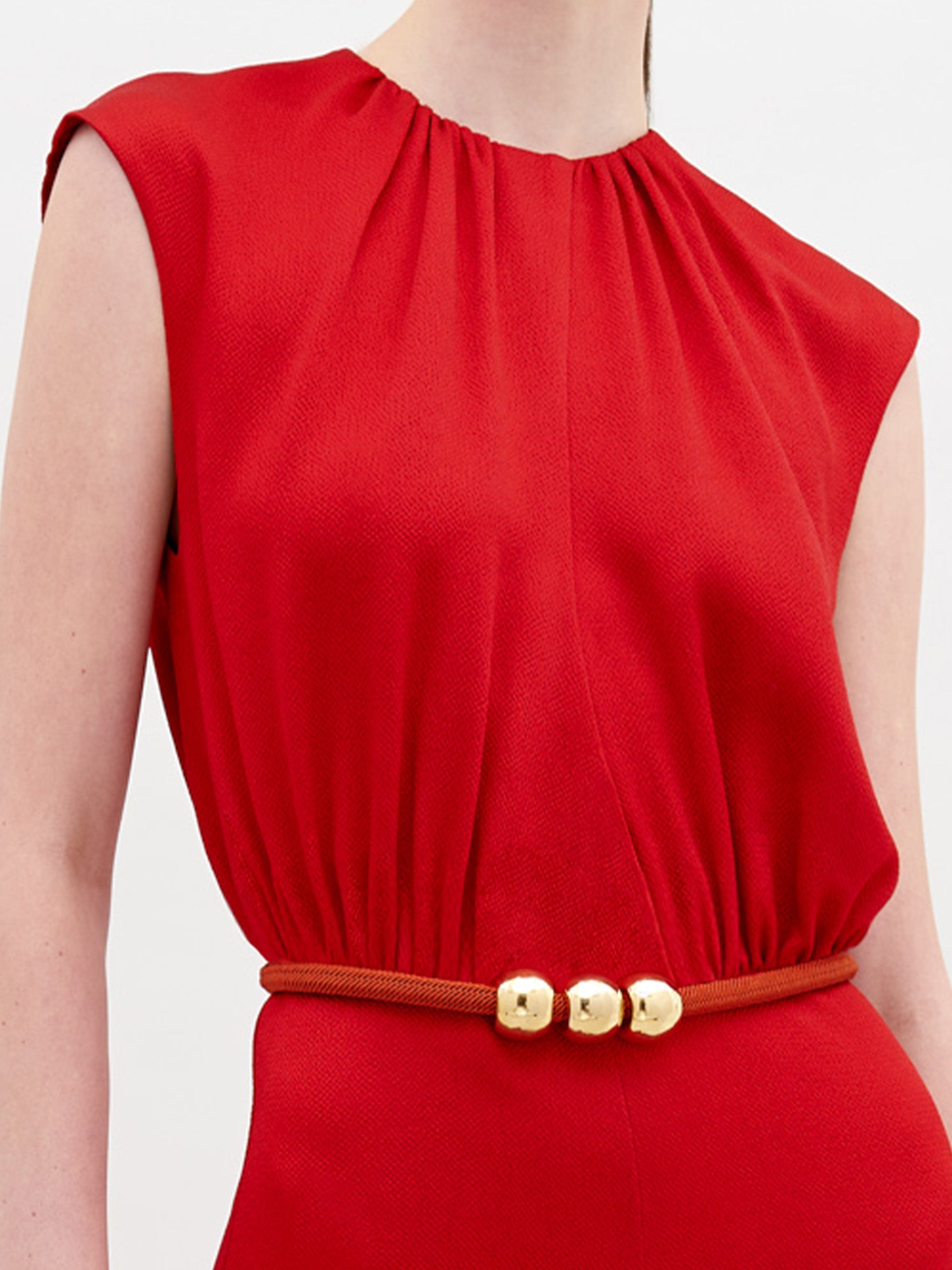 A person wearing the sleeveless Noel Jumpsuit in bright red hammered satin, featuring a gathered neckline and a slim red belt with three gold beads at the waist. The background is plain white.