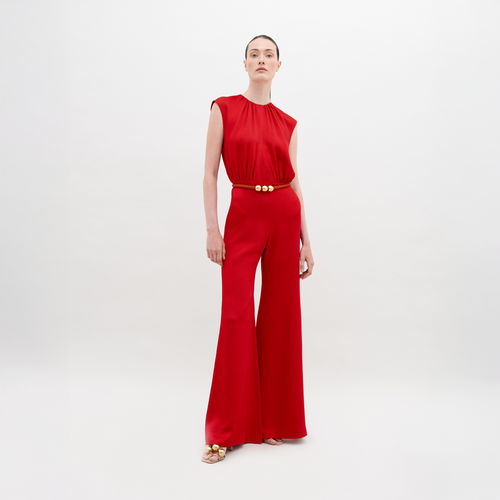 A person poses confidently against a white background, wearing the red Noel Jumpsuit by SVT DESIGNS. They have short hair and sandals, with one hand relaxed by their side and the other on their hip.
