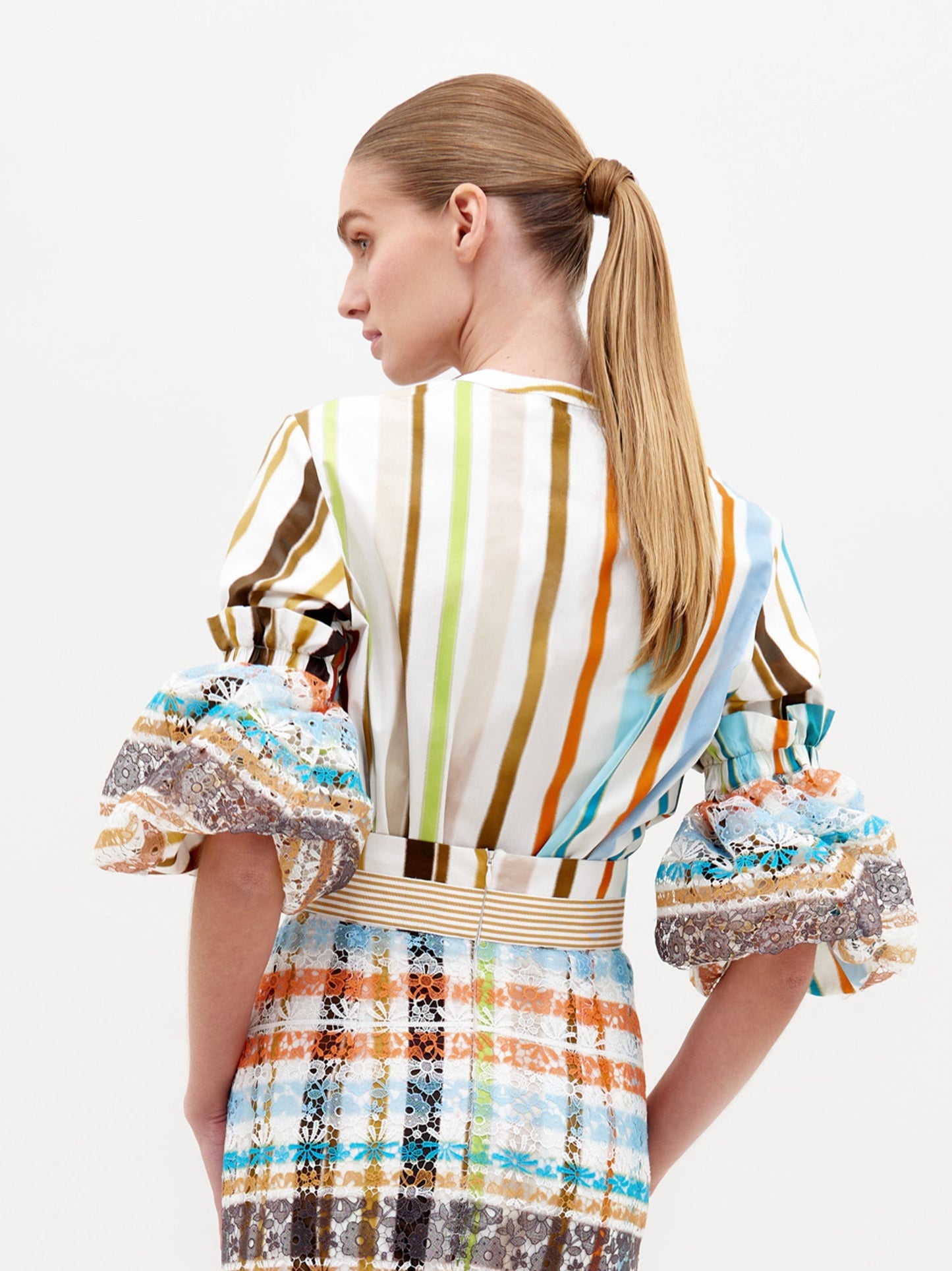 A model, dressed in the Nuru Blouse featuring multi-colored stripes and puffy sleeves, along with a patterned skirt, poses against a plain background to highlight fashion available for pre-order.