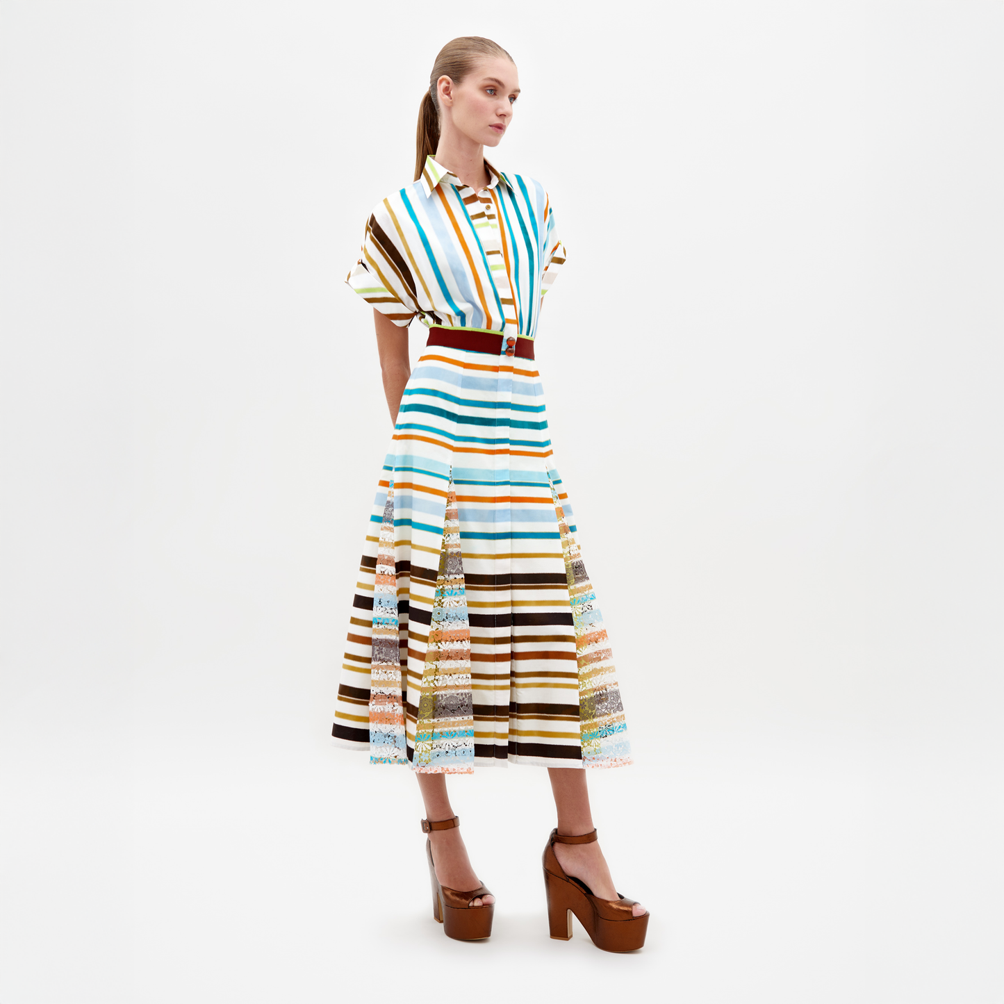 A model stands wearing the Odila Dress Multi Color Stripes paired with brown platform heels. The dress features vertical and horizontal stripes with a belted waist. This stylish ensemble, available for pre-order, will ship by early 2025. The background is plain white.