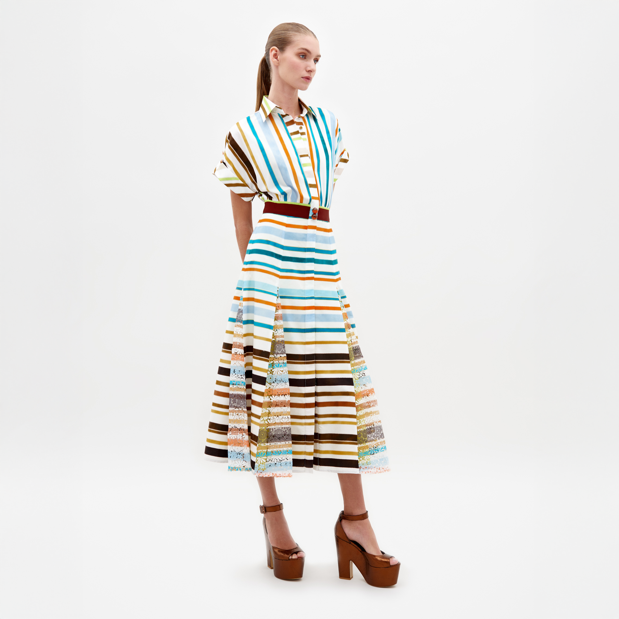A model stands wearing the Odila Dress Multi Color Stripes paired with brown platform heels. The dress features vertical and horizontal stripes with a belted waist. This stylish ensemble, available for pre-order, will ship by early 2025. The background is plain white.