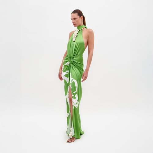 An individual in the SVT DESIGNS Olatz Dress stands against a plain white background. This sleeveless green silk dress features a high slit, floral embellishments, abstract print, high-neck halter, and knotted waist detail. They're gazing to the side.