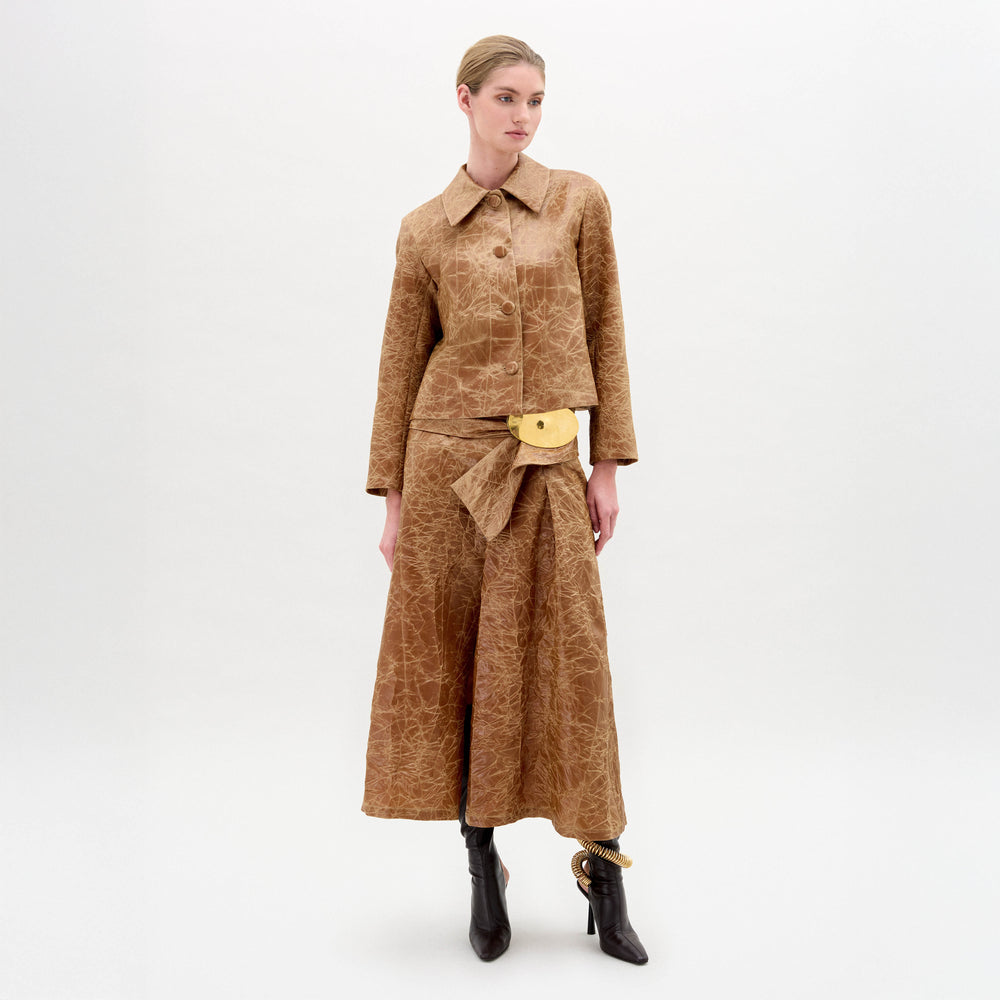 A woman stands against a plain white background wearing the SVT DESIGNS Olga Skirt, a brown textured A-line midi paired with a matching jacket, gold belt accent, and black heeled ankle boots.