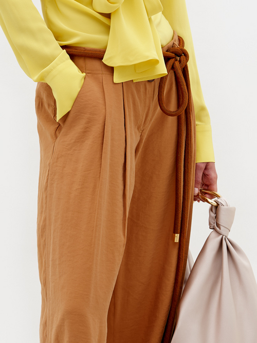 A person stands against a plain background wearing a yellow blouse, the Opal Pant Camel, and sandals, holding a large beige bag. Don't miss the chance to pre-order this outfit by February 15th, 2025.