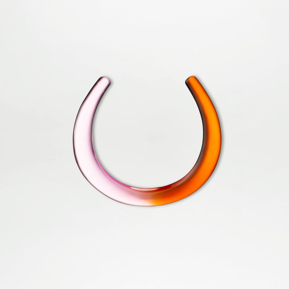 The Oba Chocker by SVT DESIGNS is a smooth, crescent-shaped glass piece made from Italian PMMA, showcasing a gradient from light pink on the left to orange on the right, set against a plain white background.