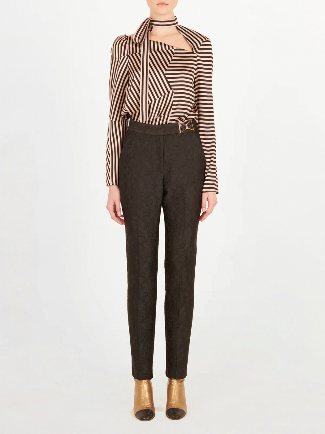 A person stands against a plain background wearing the Silvia Tcherassi Orion Pant Black, paired with a long-sleeve, black and beige geometric striped blouse and gold ankle boots. The individual's face is not visible.