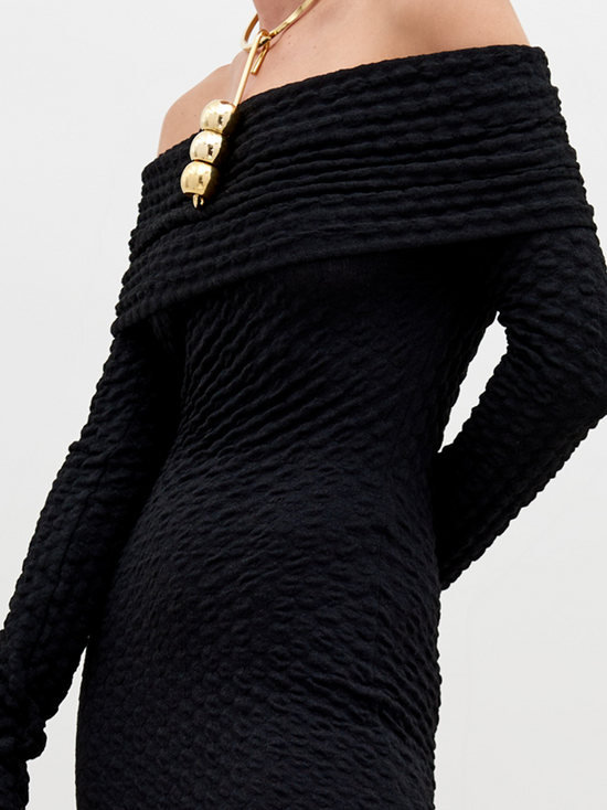 A person stands wearing a long, off-the-shoulder black textured Ozara Dress Black, holding a large black bag. They have a gold necklace and their hair is pulled back. The background is plain white, perfect for promoting the new collection available for pre-order. Mark your calendars for the ship by date: November 15th, 2024.