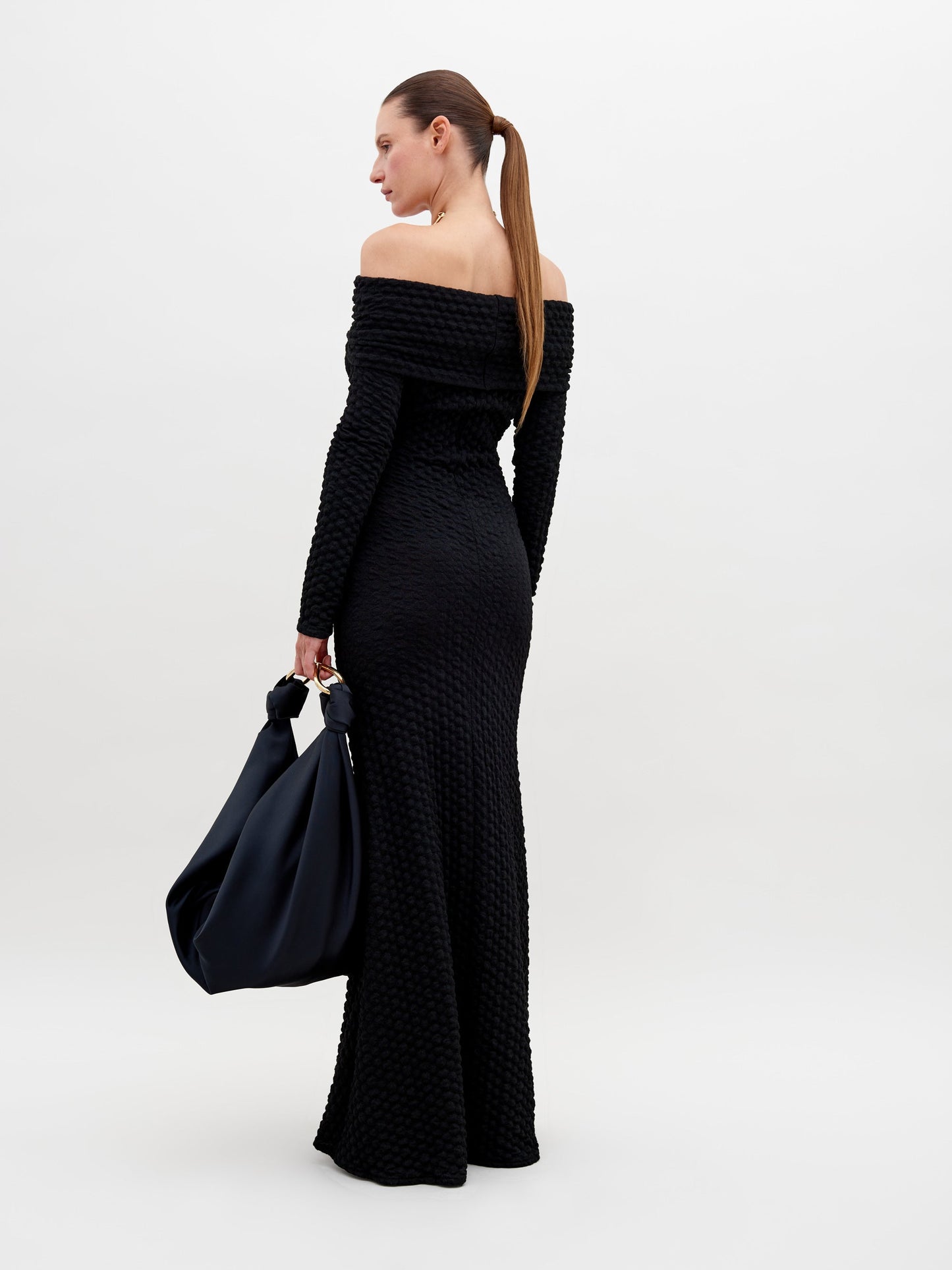 A person stands wearing a long, off-the-shoulder black textured Ozara Dress Black, holding a large black bag. They have a gold necklace and their hair is pulled back. The background is plain white, perfect for promoting the new collection available for pre-order. Mark your calendars for the ship by date: November 15th, 2024.