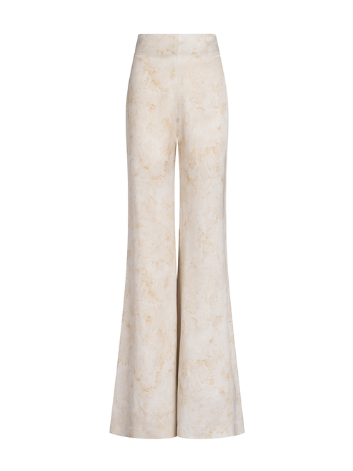 A woman stands wearing the Palermo Pant in Cream Abstract Print, a strapless jumpsuit featuring a light-colored fabric and a floral design on the top, accessorized with a small clutch against a plain blue background. Pre-order now for August shipping dates and elevate your summer wardrobe effortlessly.