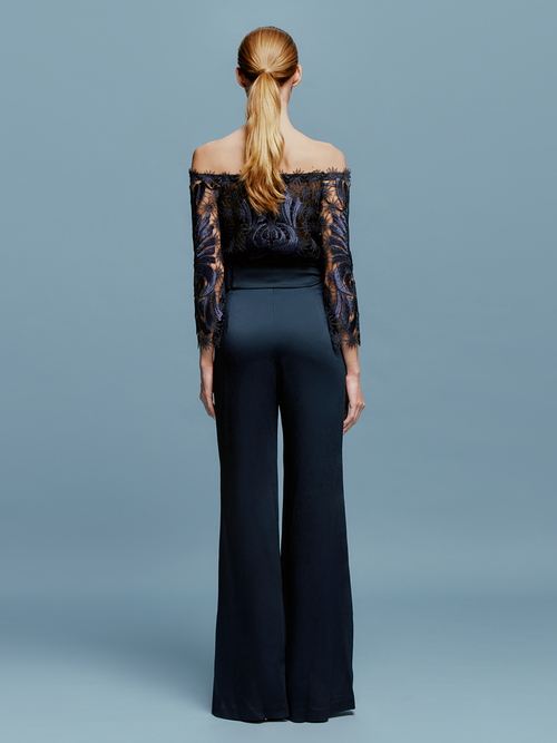 Woman standing against a blue background, wearing an off-the-shoulder navy patterned top and the Palermo Pant Navy, ready to set sail on September 2nd.