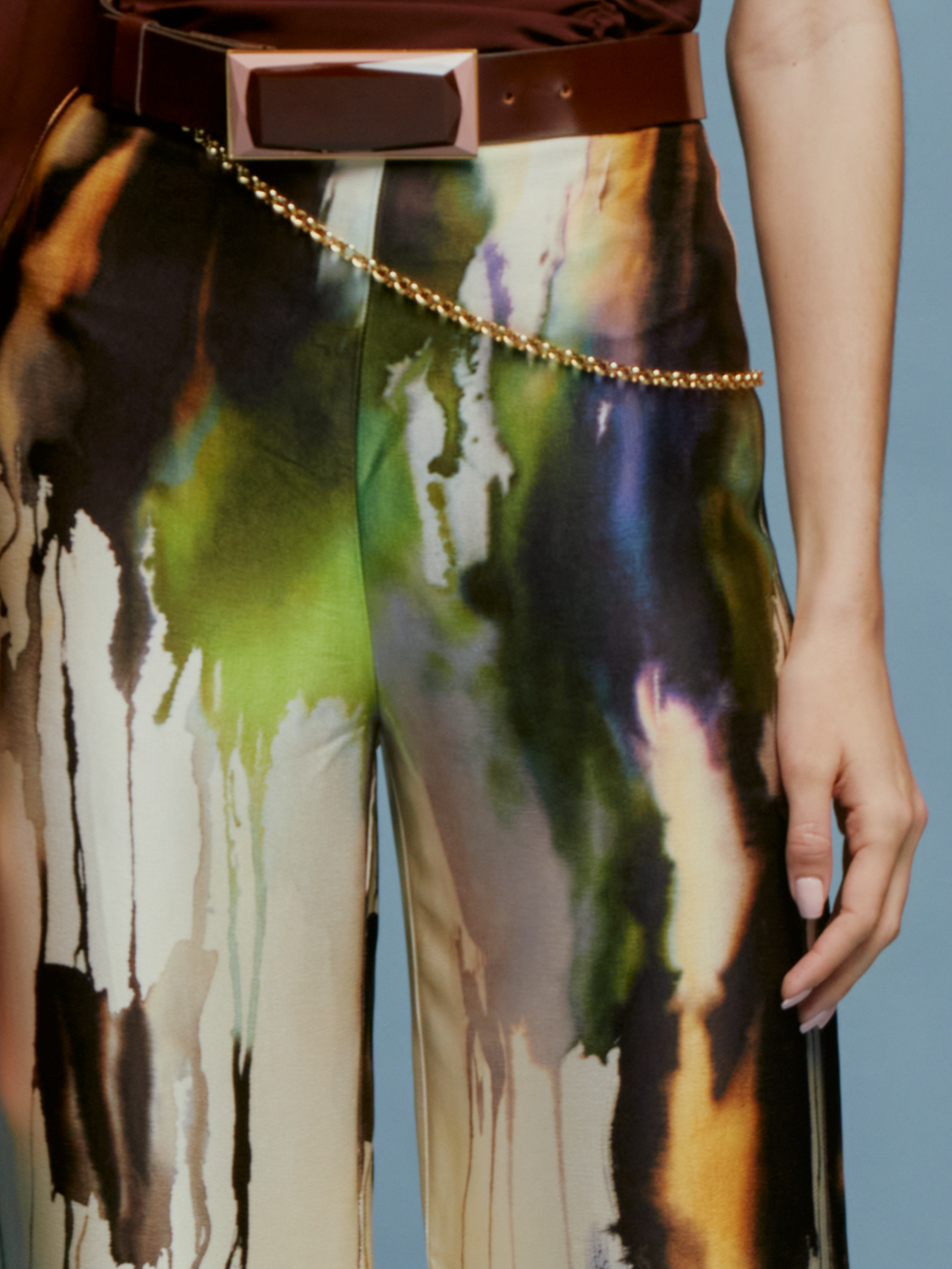 A female model wearing a one-shoulder brown top and wide-leg Palermo Pant in Olive Watercolor stands against a plain blue background, ready to set sail on her fashion journey.