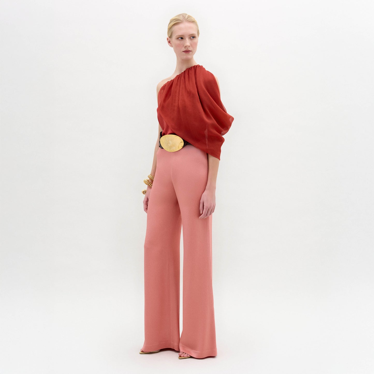 A woman stands in high-waisted pink Palermo Pants, crafted from luxurious Italian fabric, paired with a red one-shoulder top, gold belt, gold bracelets, and open-toe shoes against a white background.