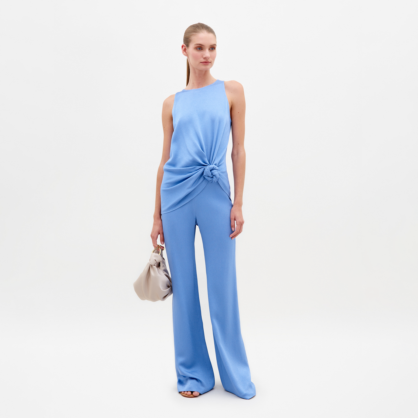 A woman wearing a sleeveless, periwinkle top with a knot at the waist and matching Palermo Pant in Periwinkle stands holding a beige handbag against a plain white background. Pre-order now for delivery by February 15th, 2025!
