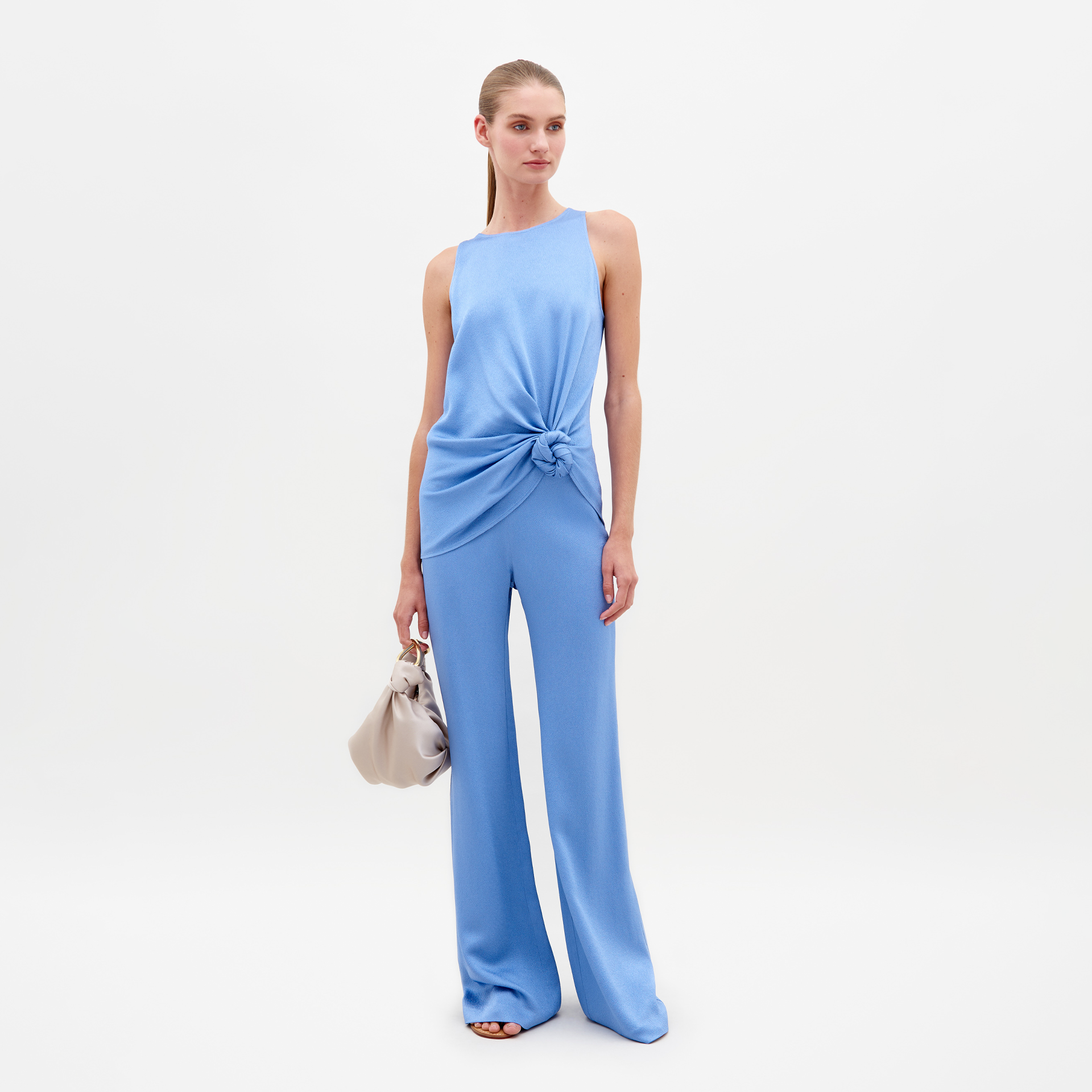 A woman wearing a sleeveless, periwinkle top with a knot at the waist and matching Palermo Pant in Periwinkle stands holding a beige handbag against a plain white background. Pre-order now for delivery by February 15th, 2025!