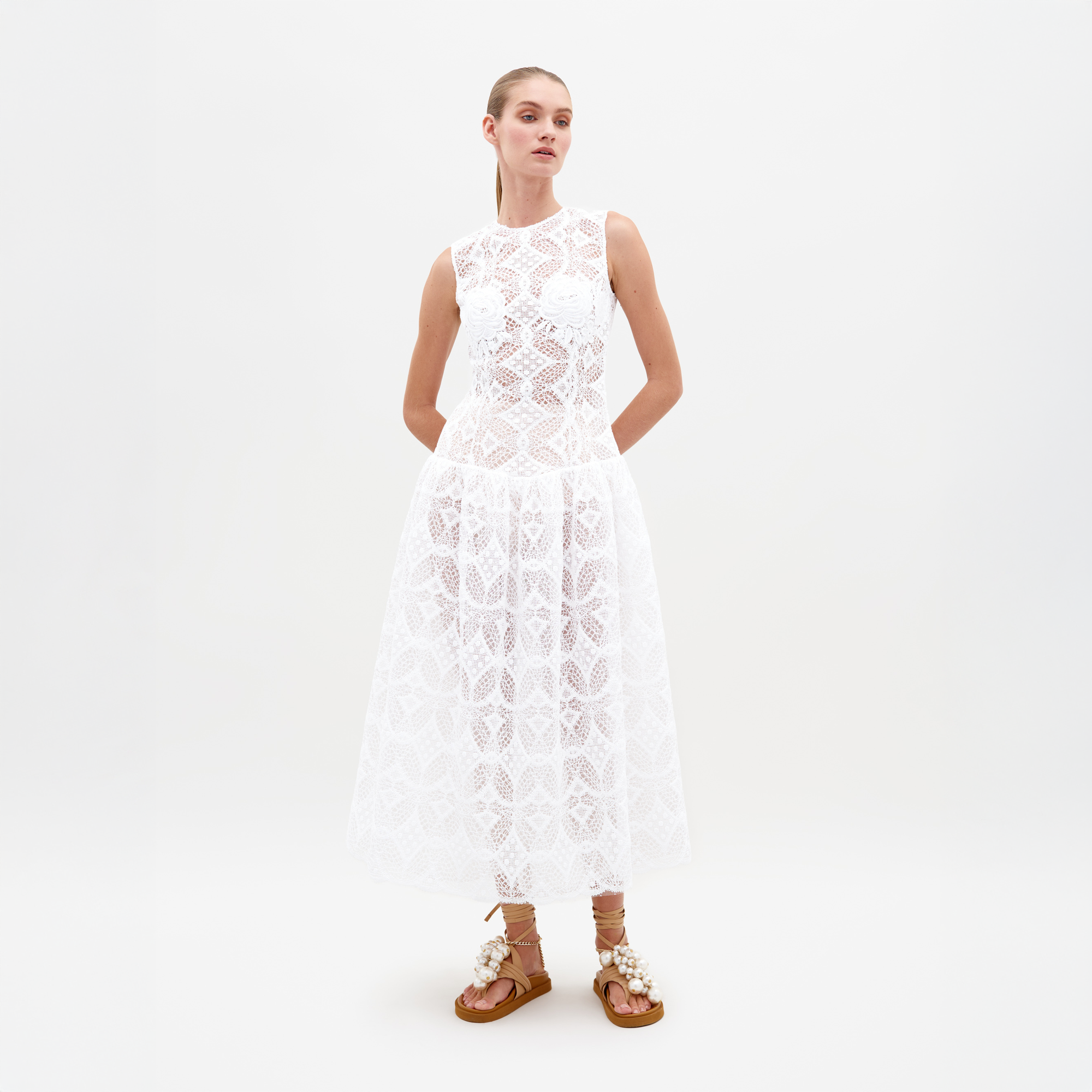 A person stands in the Paola Dress White, featuring a sleeveless lace design with a flared skirt, complemented by brown lace-up sandals. This item is available for pre-order and will ship by February 15th, 2025. The background is plain white.
