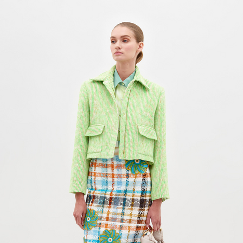 A person is wearing the Perla Jacket by SVT DESIGNS, tailored in light green tweed with two front pockets, over a multi-colored plaid skirt featuring floral patterns. A light blue shirt with a traditional collar peeks out from under the jacket against a plain white background.