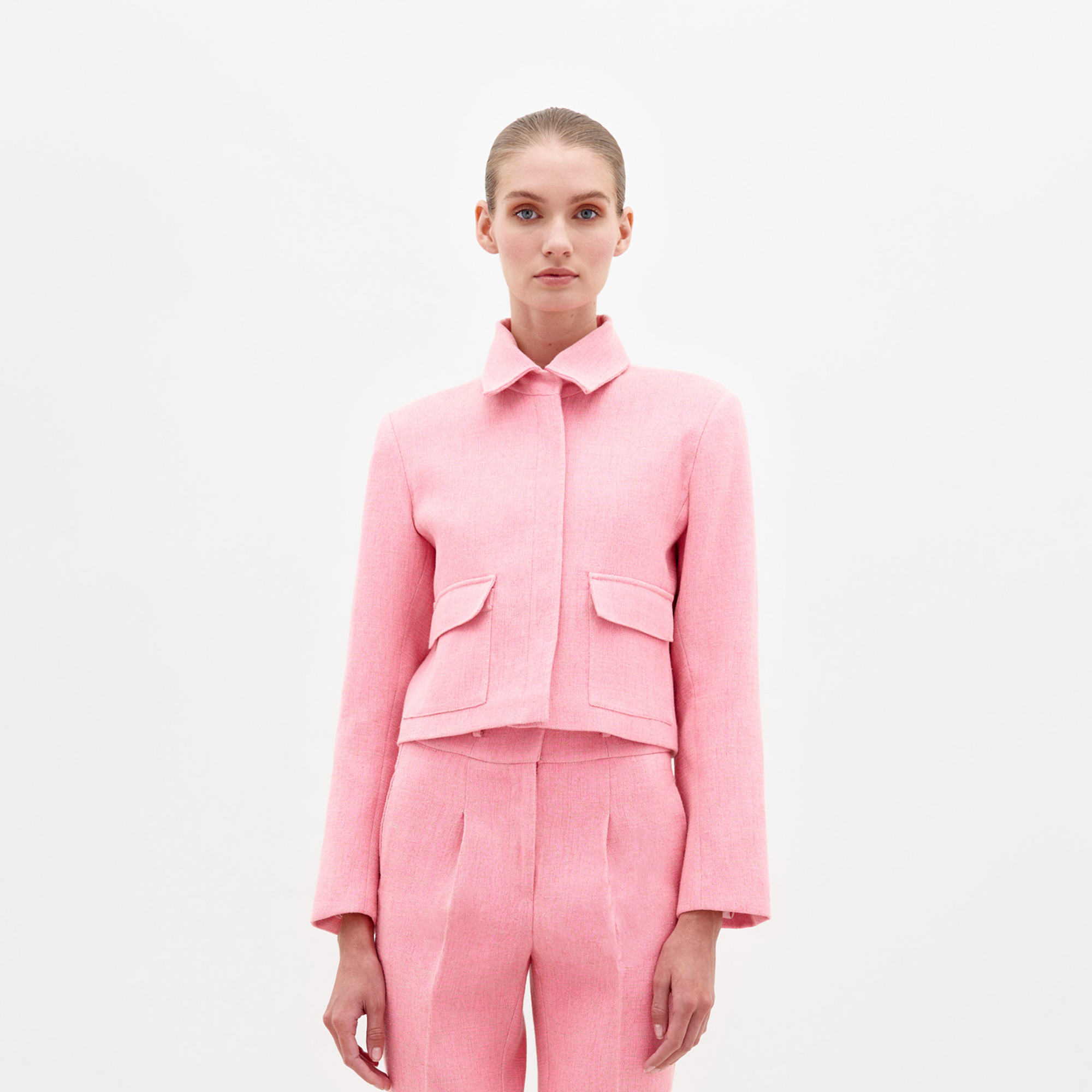 Against a plain white background, someone wears the SVT DESIGNS Perla Jacket, a pink suit made from exquisite Italian fabric. The expertly tailored collared jacket features two front flap pockets, and their light hair is pulled back neatly, adding elegance to the ensemble.