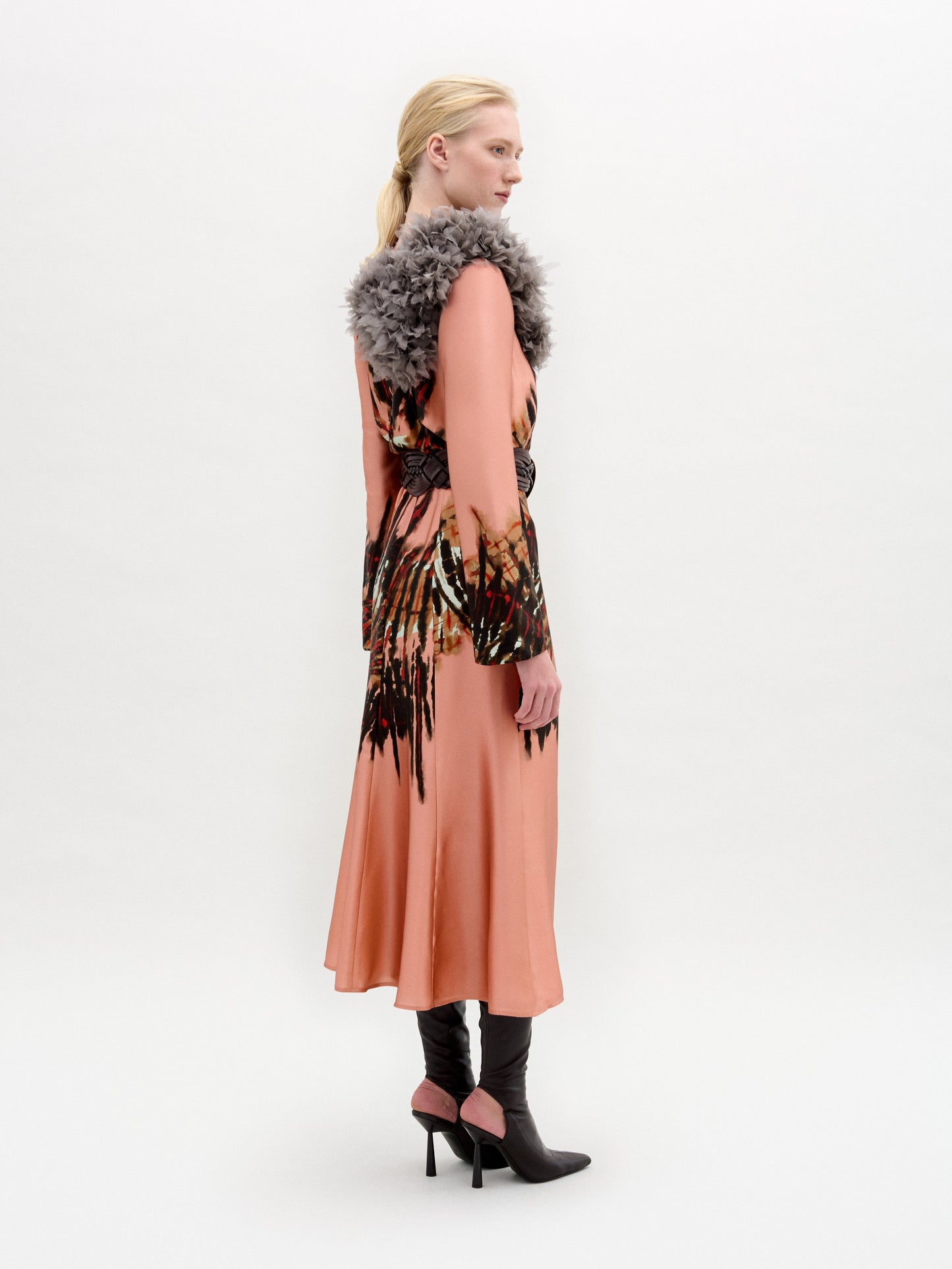 A person stands in profile wearing the Pesaro Dress—a peach silk midi with a large feathered design, wide belt with round buckle, gray fur collar, and black heeled boots—set against a plain background.